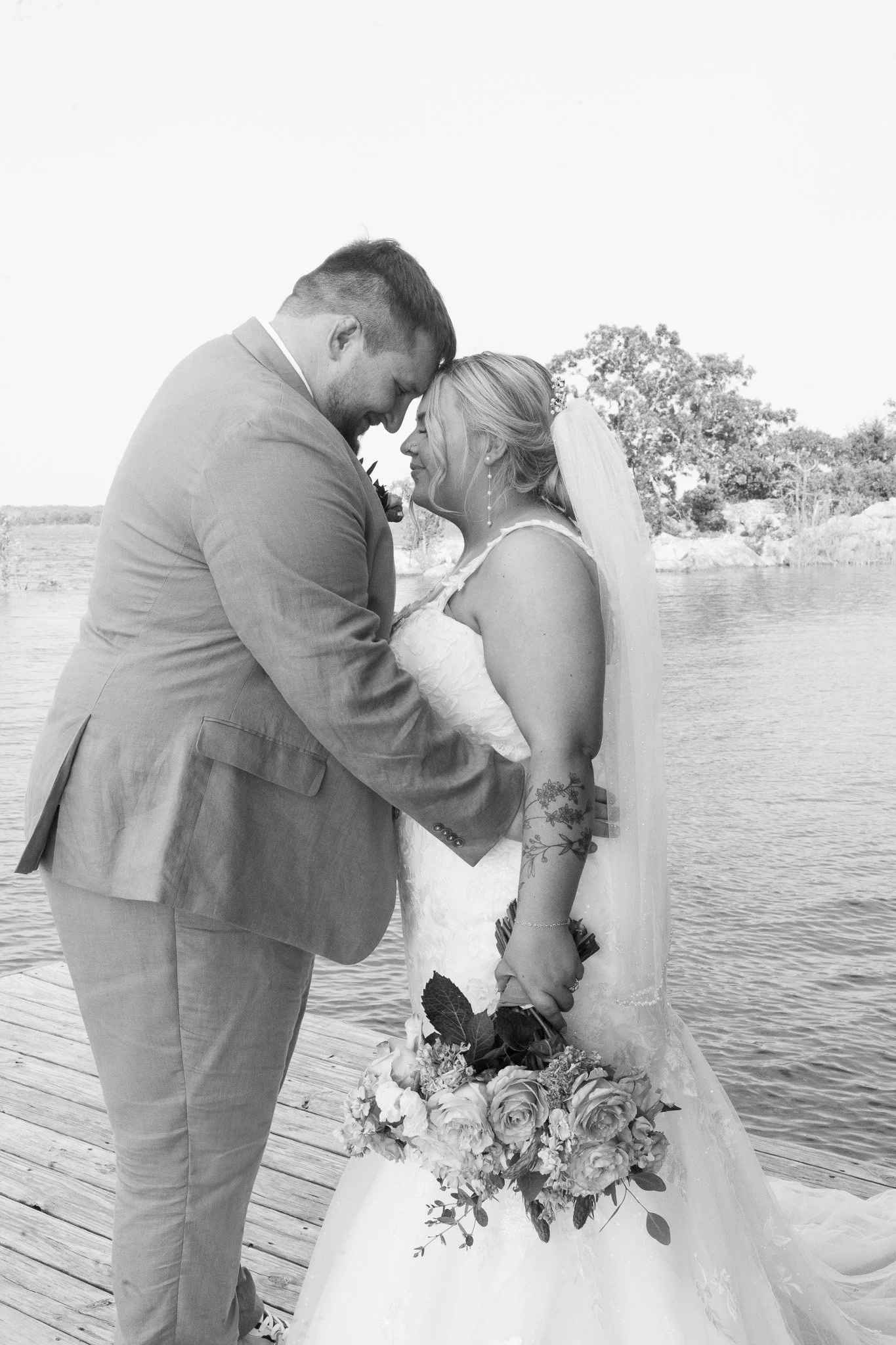 A black and white photo of a bride and groom on a dock, holding hands and about to kiss by a lake, with trees in the background. The bride holds a bouquet of flowers, and the groom wears a suit.