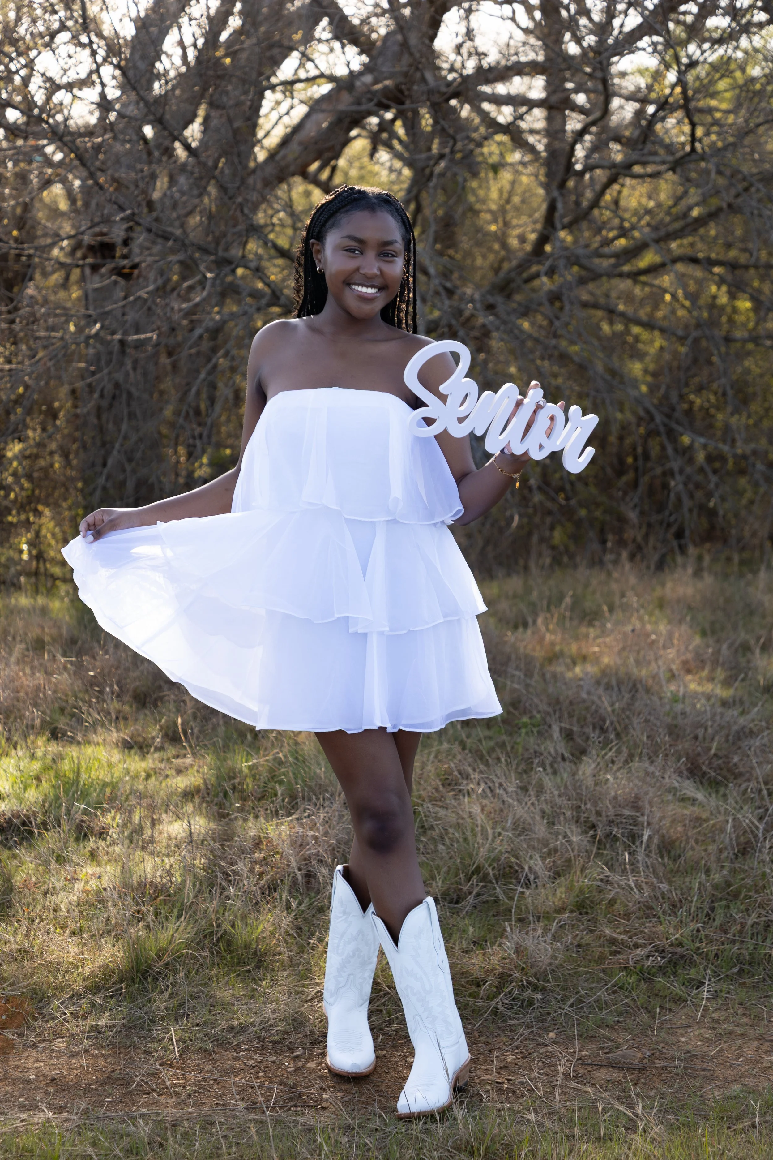 A young woman in a white, strapless, layered dress and cowboy boots standing in a grassy outdoor area with bare trees in the background. She is smiling and holding a sign that says 'Senior'.