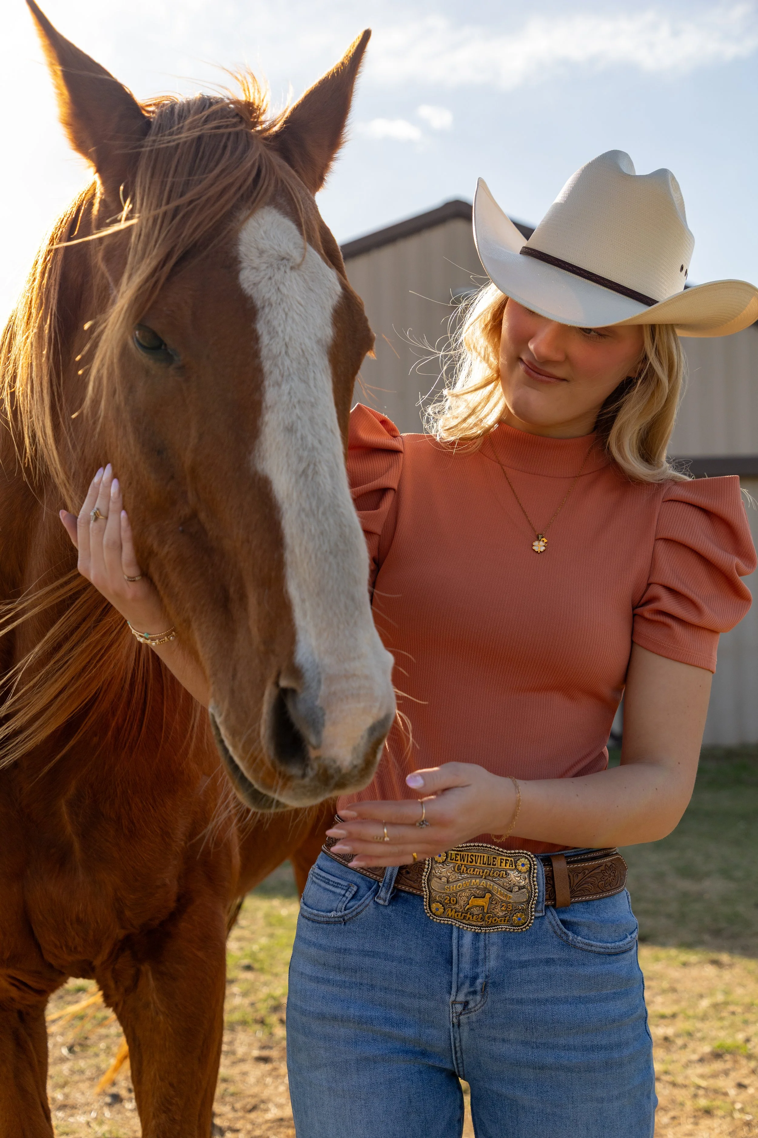 Young woman wearing a cowboy hat and a peach-colored top standing beside a chestnut horse with a white blaze, outdoors during golden hour.