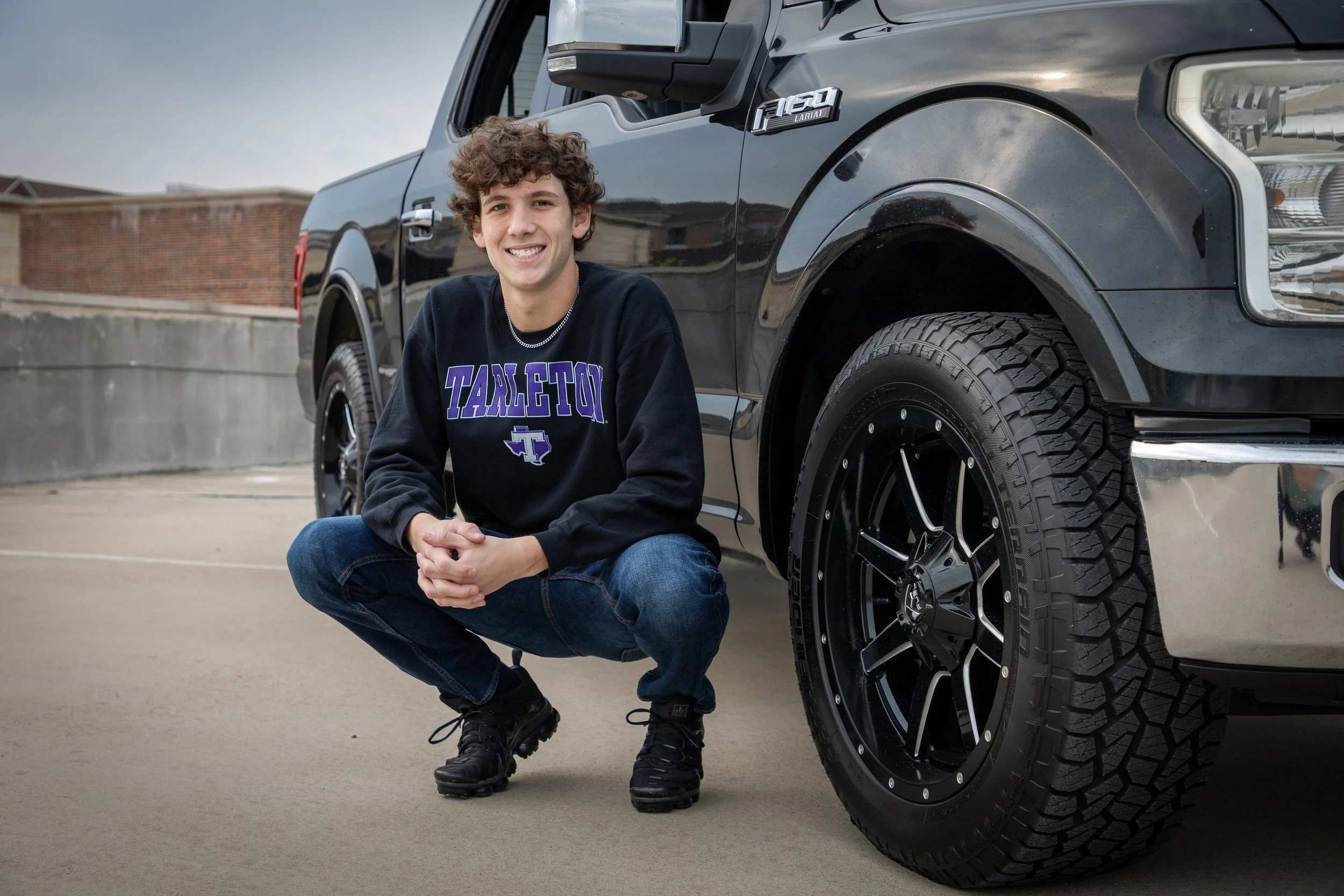 A young man crouching next to a black pickup truck on a rooftop parking lot, smiling at the camera. He is wearing a black sweatshirt with purple lettering that reads 'TARKETON' and jeans.