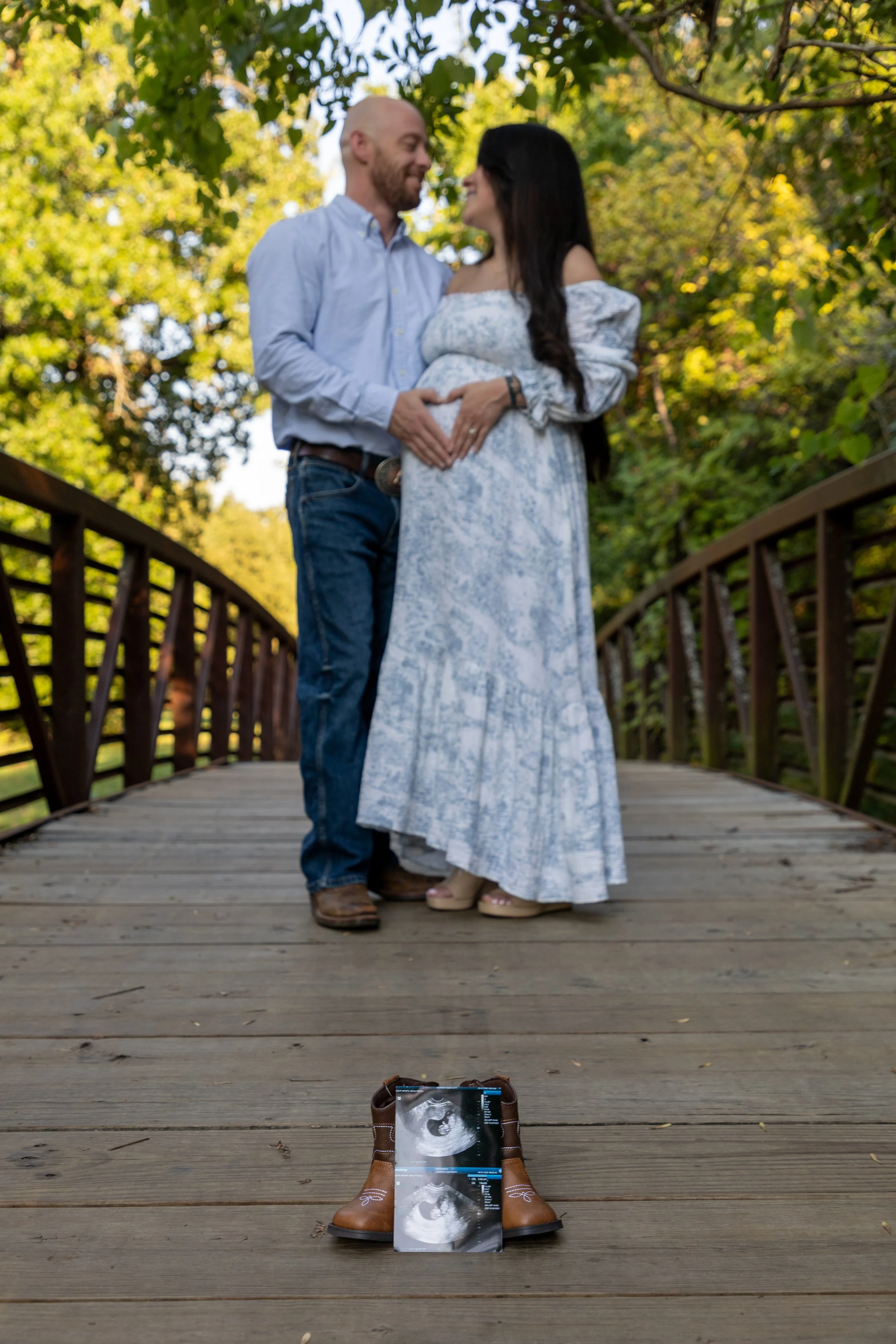A couple stands on a wooden bridge, facing each other, with a sonogram on the ground in front of them and the woman is pregnant.