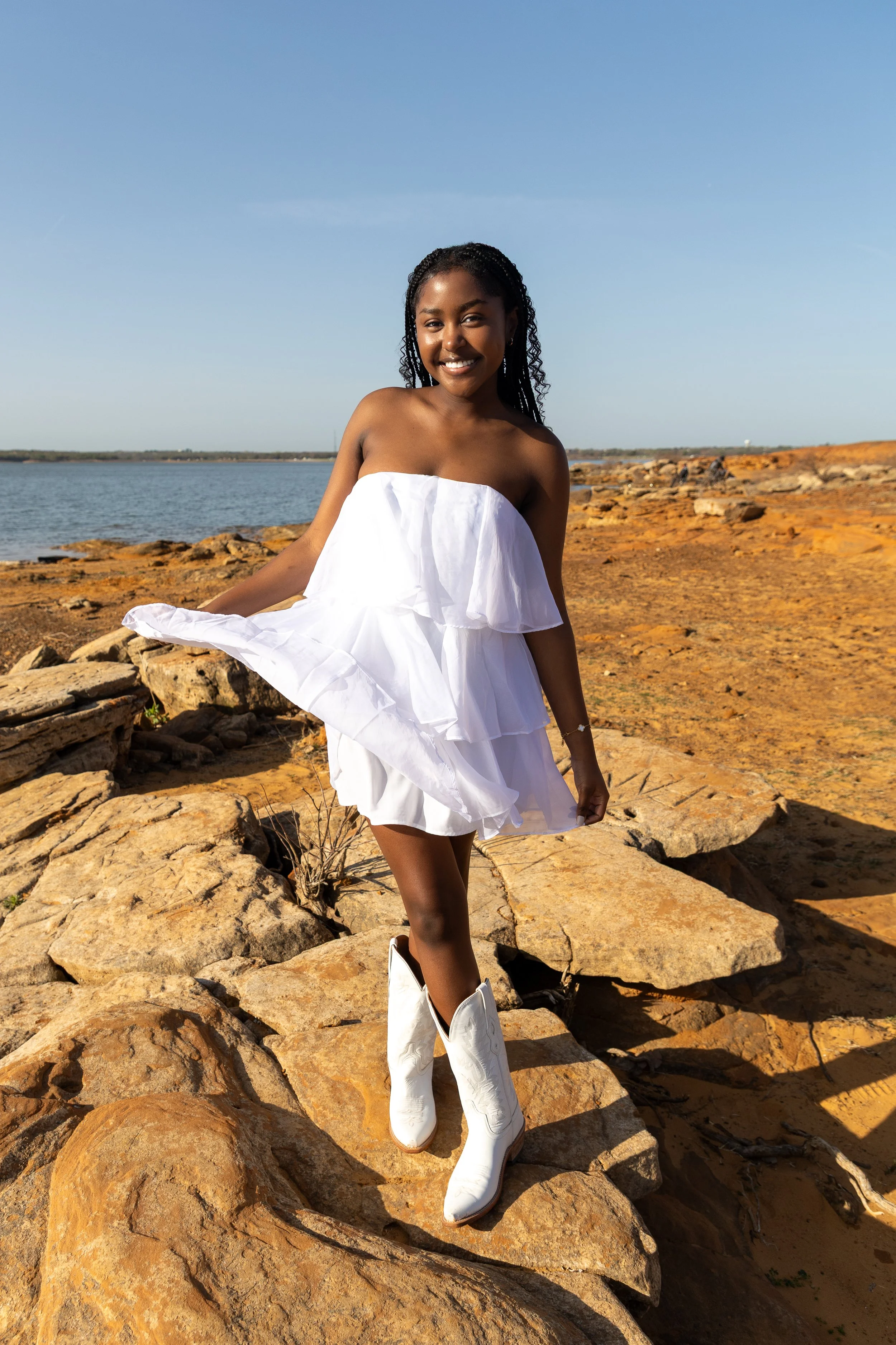 A woman standing on rocks near a body of water, wearing a white strapless dress and white cowboy boots, smiling at the camera in a sunny outdoor setting.