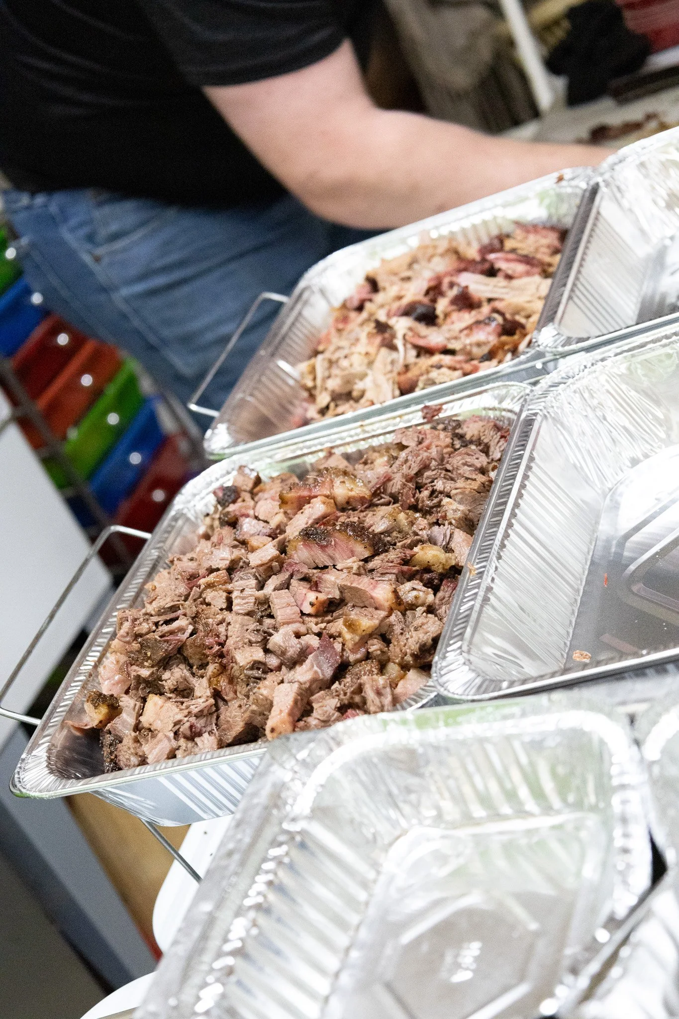Two aluminum trays filled with chopped cooked meat, likely beef or pork, placed on a metal stand, with a person in a black shirt and jeans in the background.