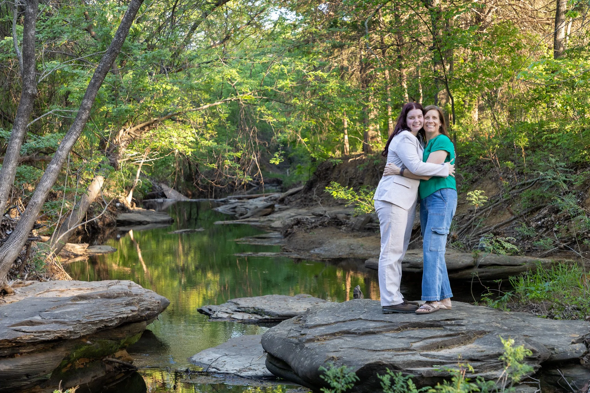 Two women hugging on a large rock in a forested creek. Sunlight filters through green trees.