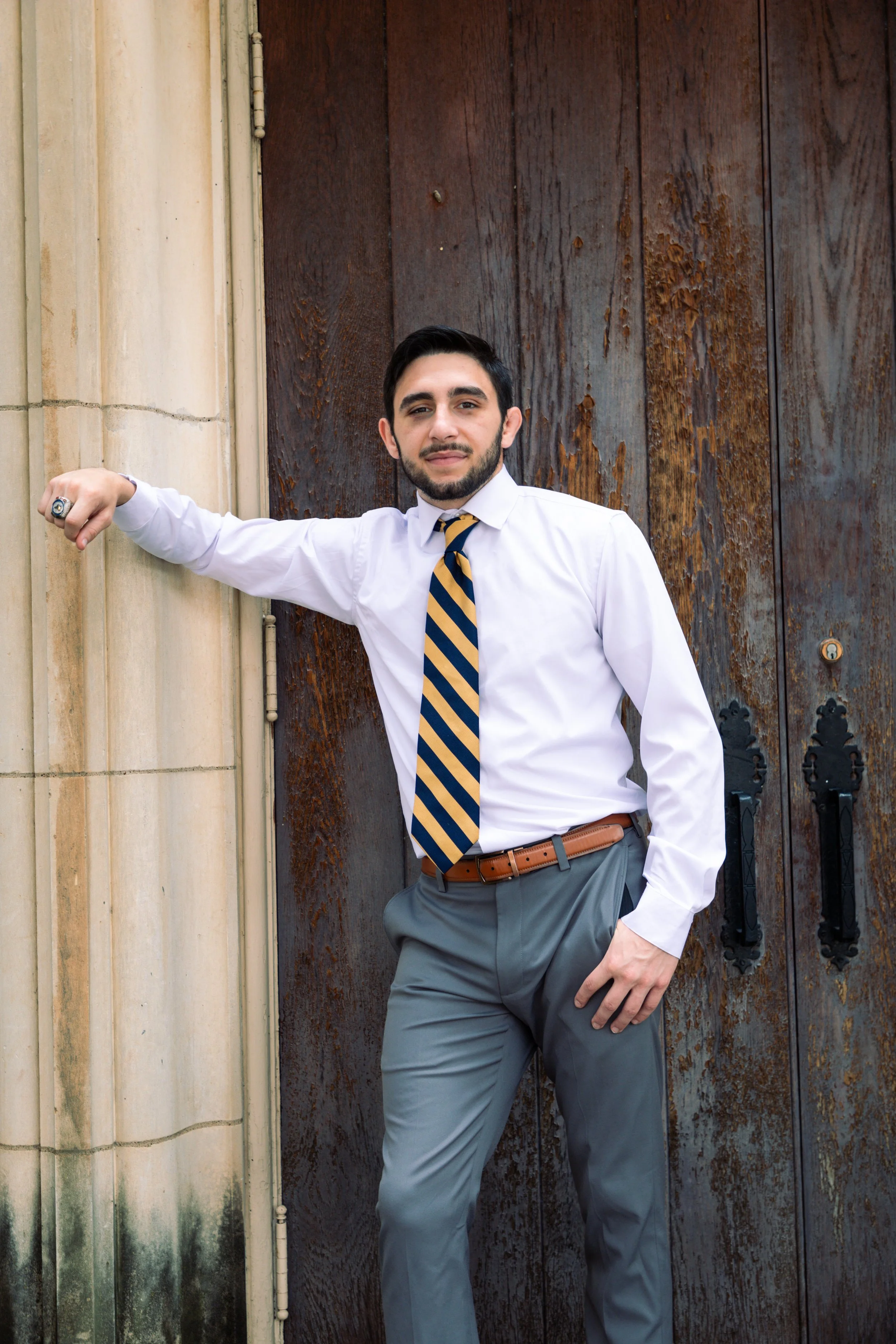A man in business attire standing against a wooden door and concrete wall, with one arm resting on the wall and the other in his pocket.