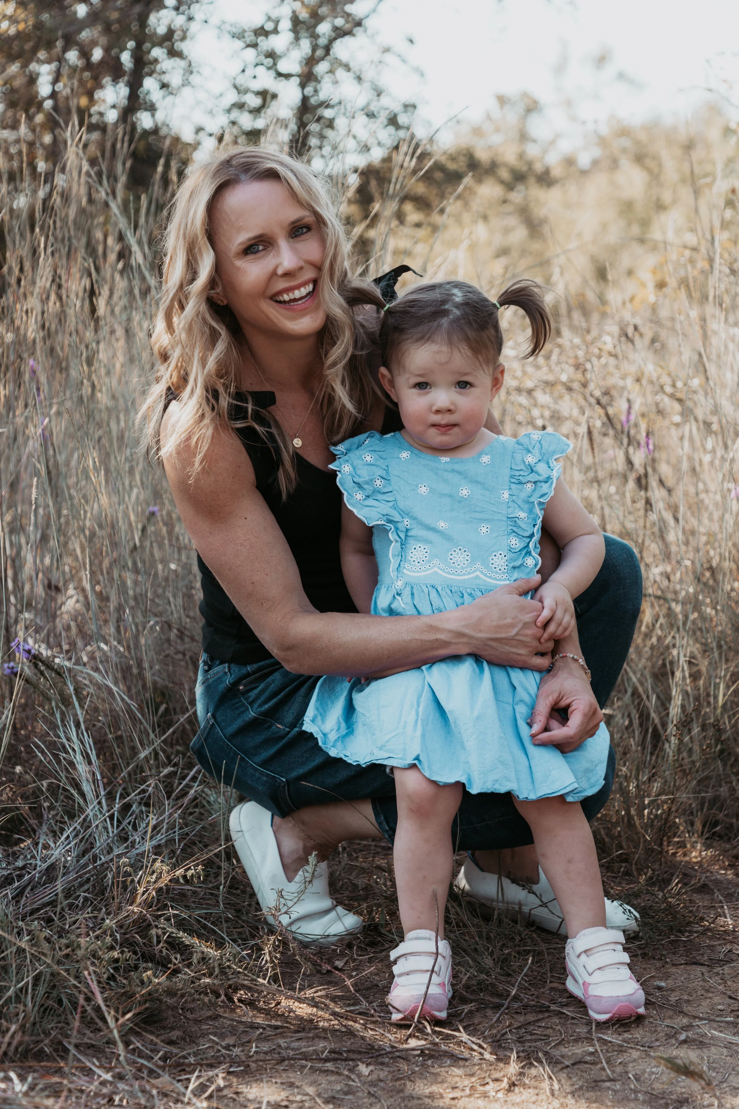 A woman with blonde wavy hair and a young girl with pigtails pose together outdoors in a field of tall grass and wildflowers during daytime. The woman is smiling and wearing a black top and ripped jeans, while the girl in a light blue dress has a neu