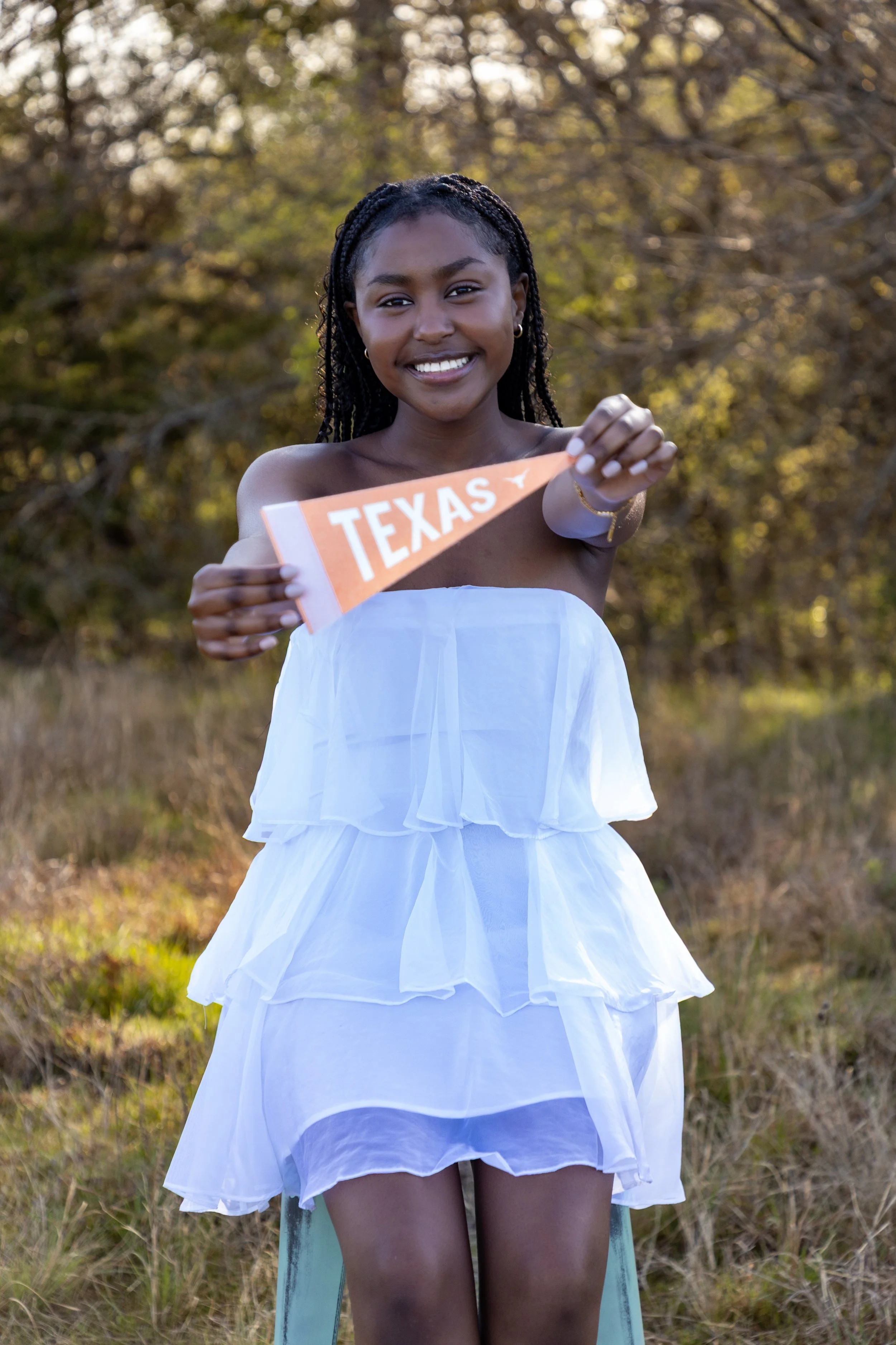 A young woman smiling outdoors holding an orange Texas pennant flag, wearing a white layered dress and standing in a grassy area with trees in the background.