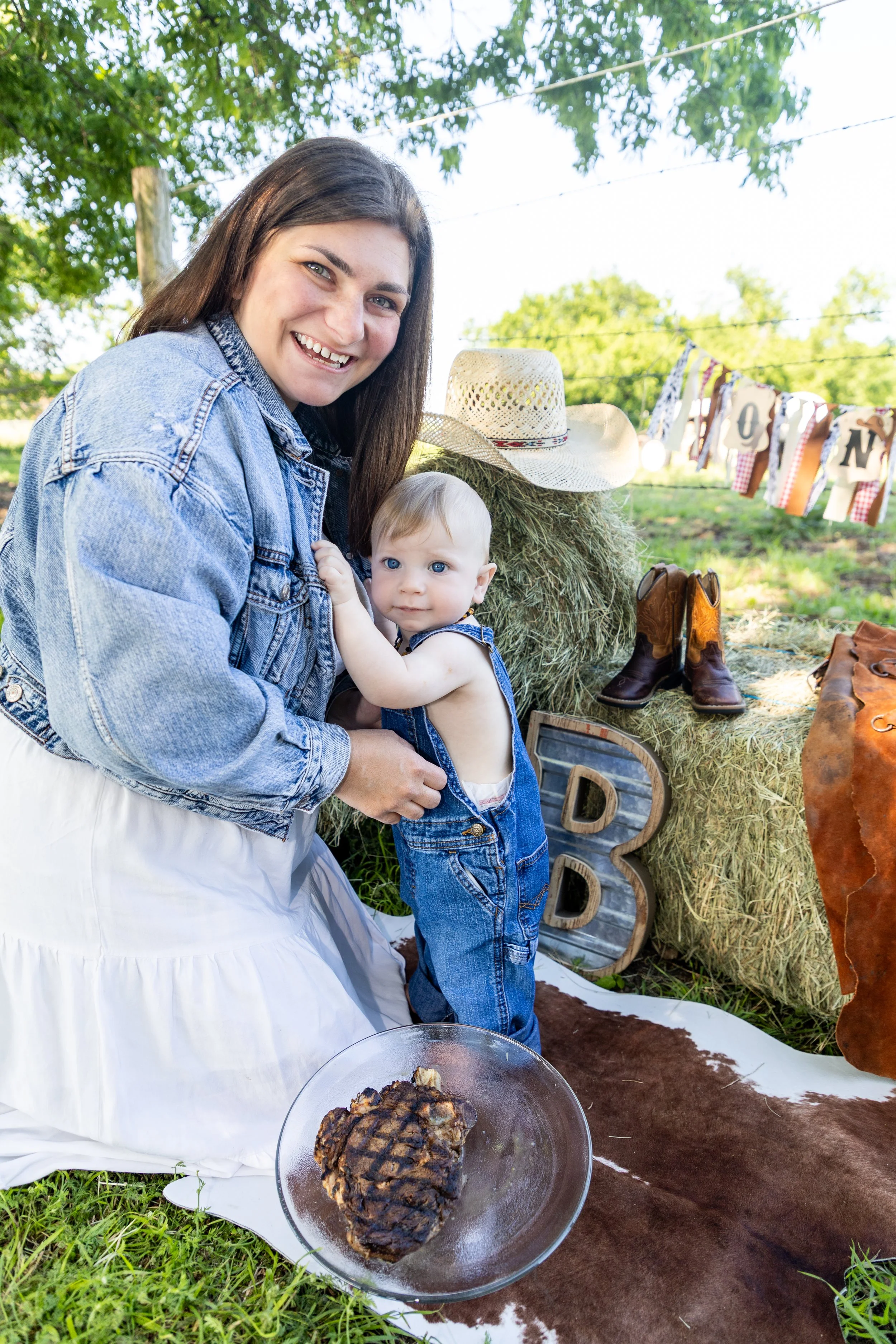 A woman and a baby in denim overalls pose in a rural outdoor setting with hay bales, cowboy boots, a straw hat, decorative letters, and a fire-grilled steak.