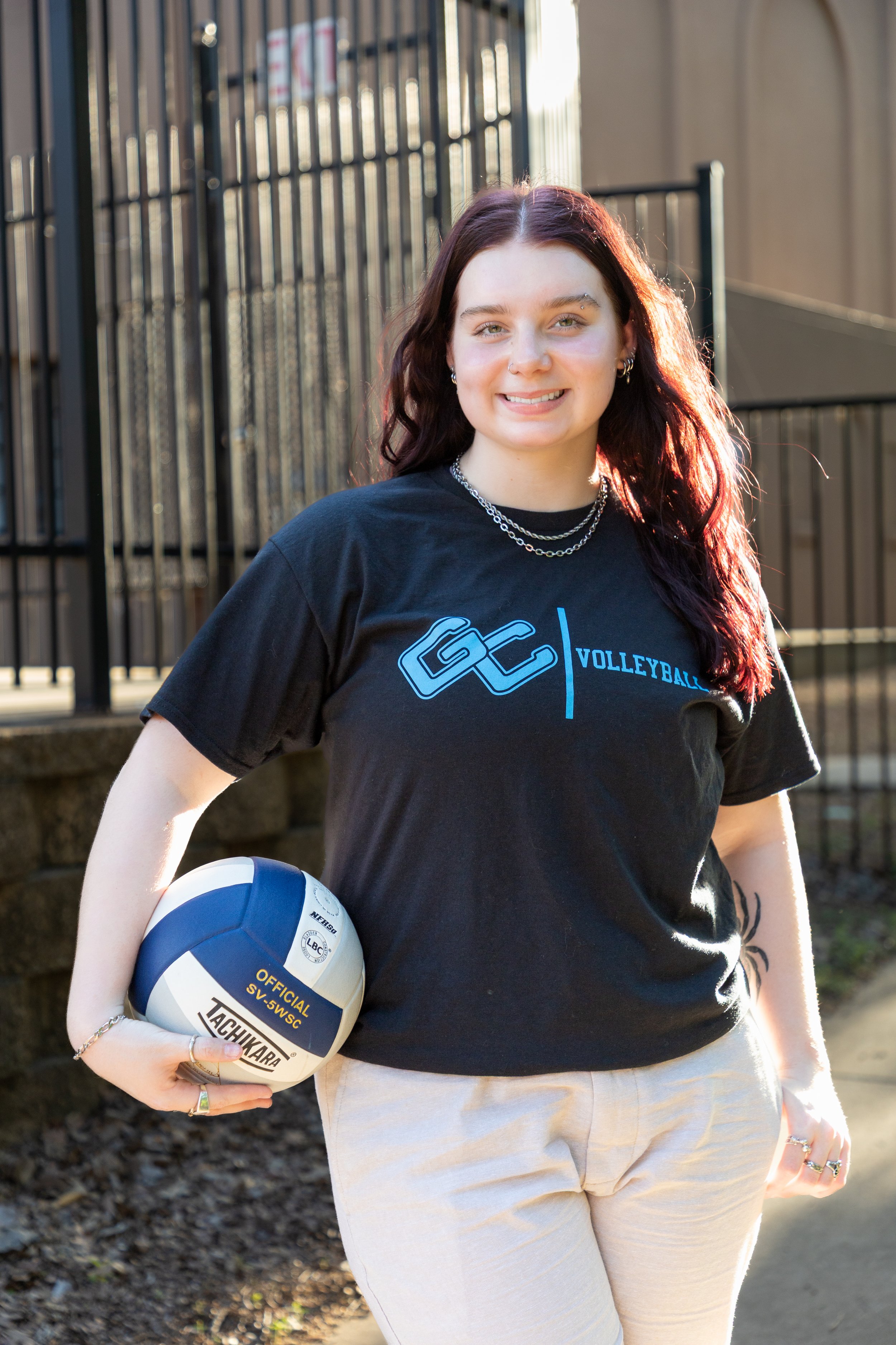 A young woman with long dark red hair smiling outdoors, holding a white and blue volleyball, wearing a black t-shirt with a volleyball logo, and standing next to a black metal fence.