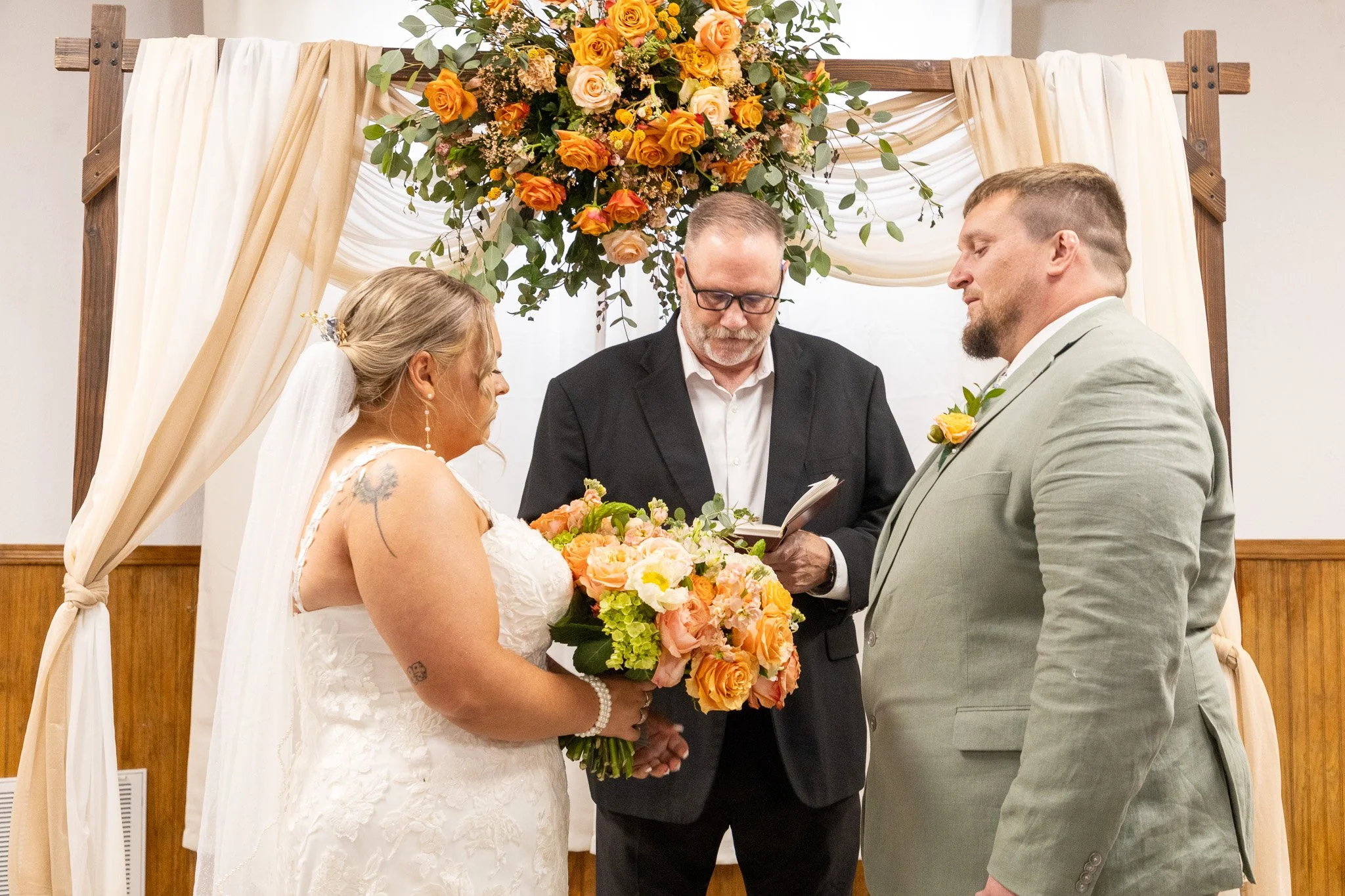 A bride and groom stand in front of an officiant, exchanging vows during a wedding ceremony, with a floral arrangement and draped fabric backdrop behind them.