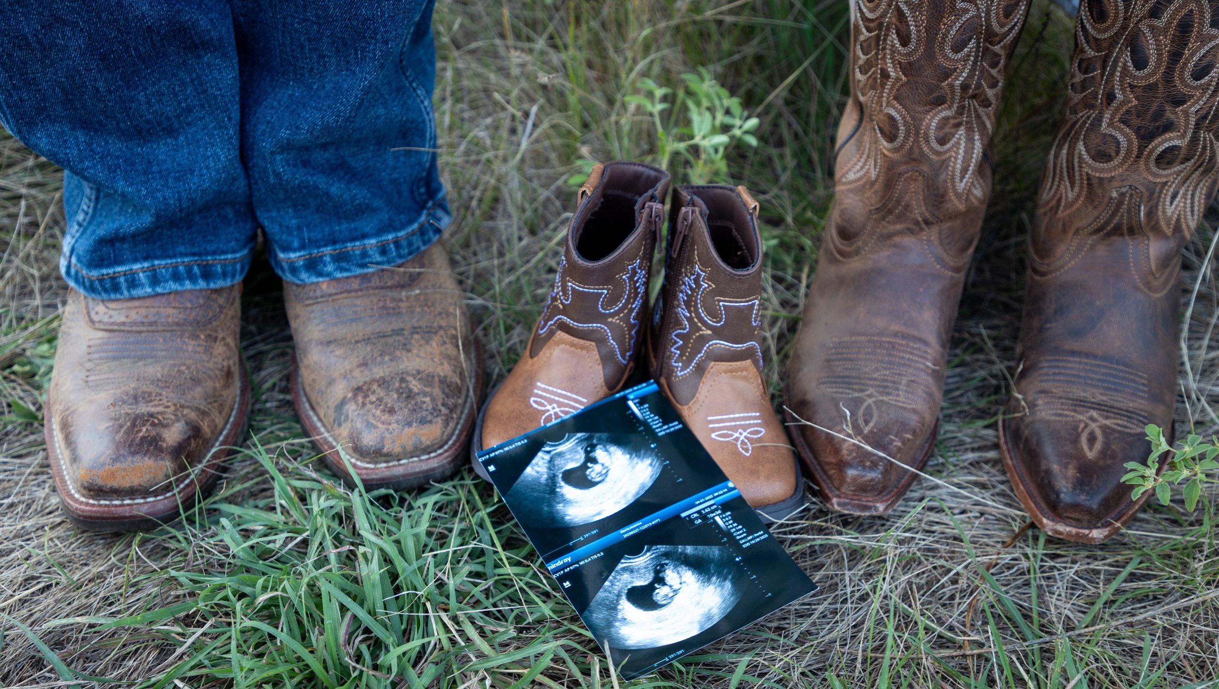 Three pairs of cowboy boots and two ultrasound photos on grass, two of the boots are brown with embroidery, one pair is darker brown with a more worn appearance.