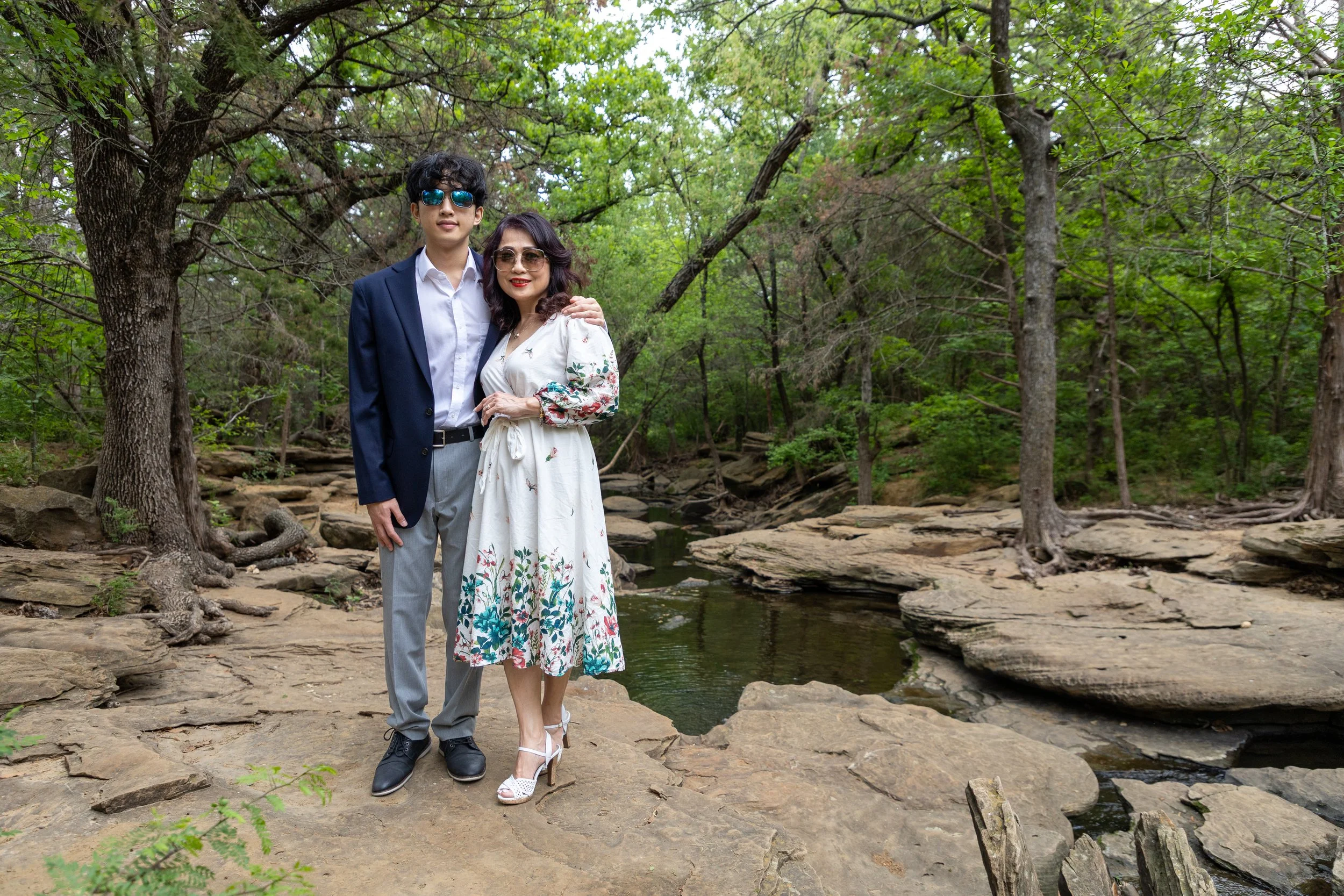 A young man in sunglasses and a woman in sunglasses and a white floral dress standing on a rocky area near a creek in a green forest.