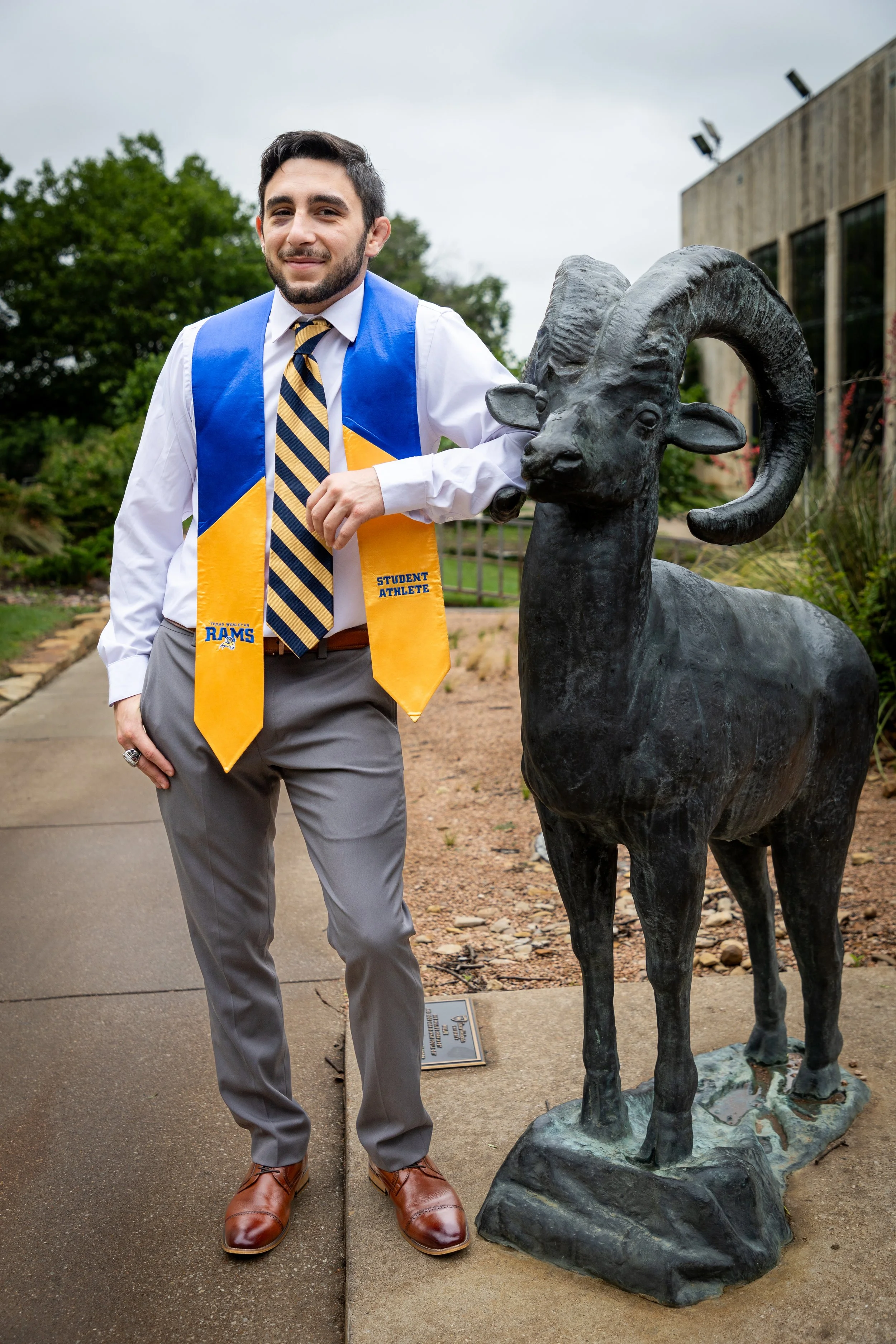 Young man in dress shirt, tie, and gray pants standing on sidewalk next to bronze ram statue, leaves and trees in background.
