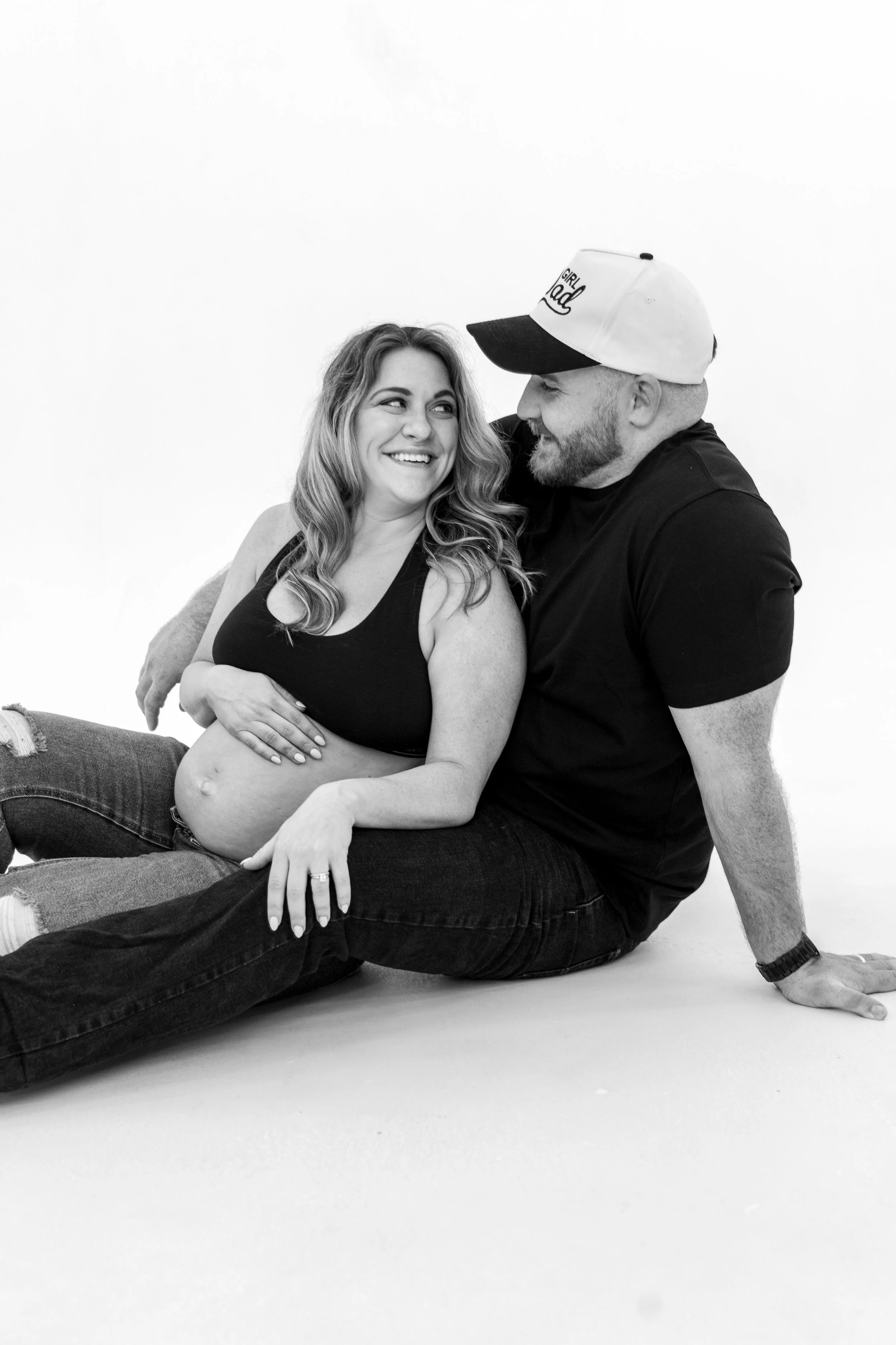 A smiling pregnant woman with wavy hair and a sleeveless top sitting on the lap of a man with a beard, wearing a baseball cap with the words "Girl Dad," in a casual setting with a plain white background.