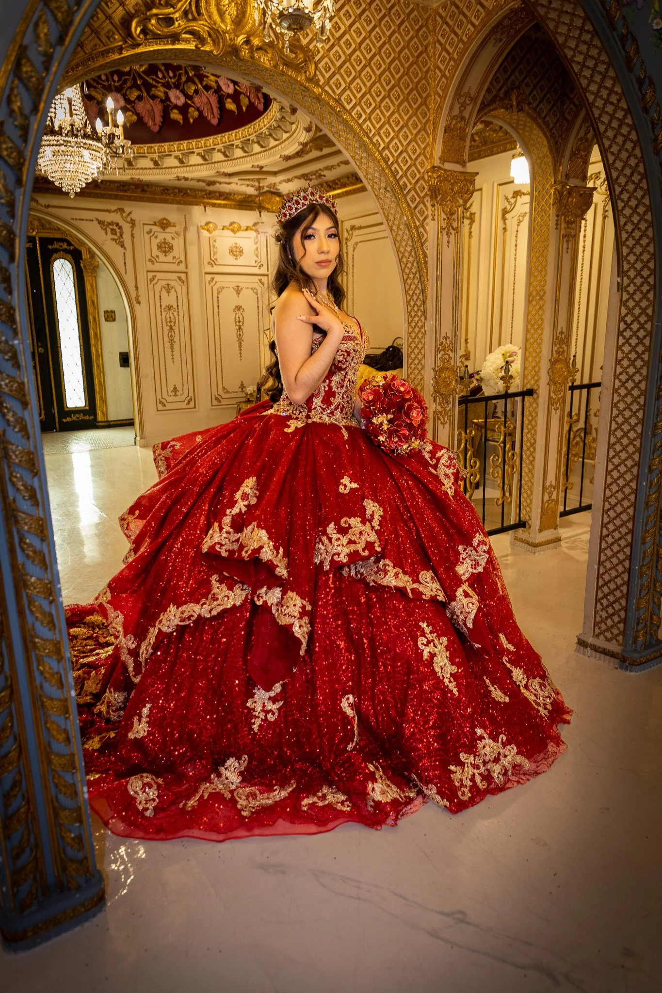 A woman dressed in an elaborate red and gold wedding gown with intricate embroidery, standing in a luxurious room with ornate gold and white decor, chandeliers, and arches.