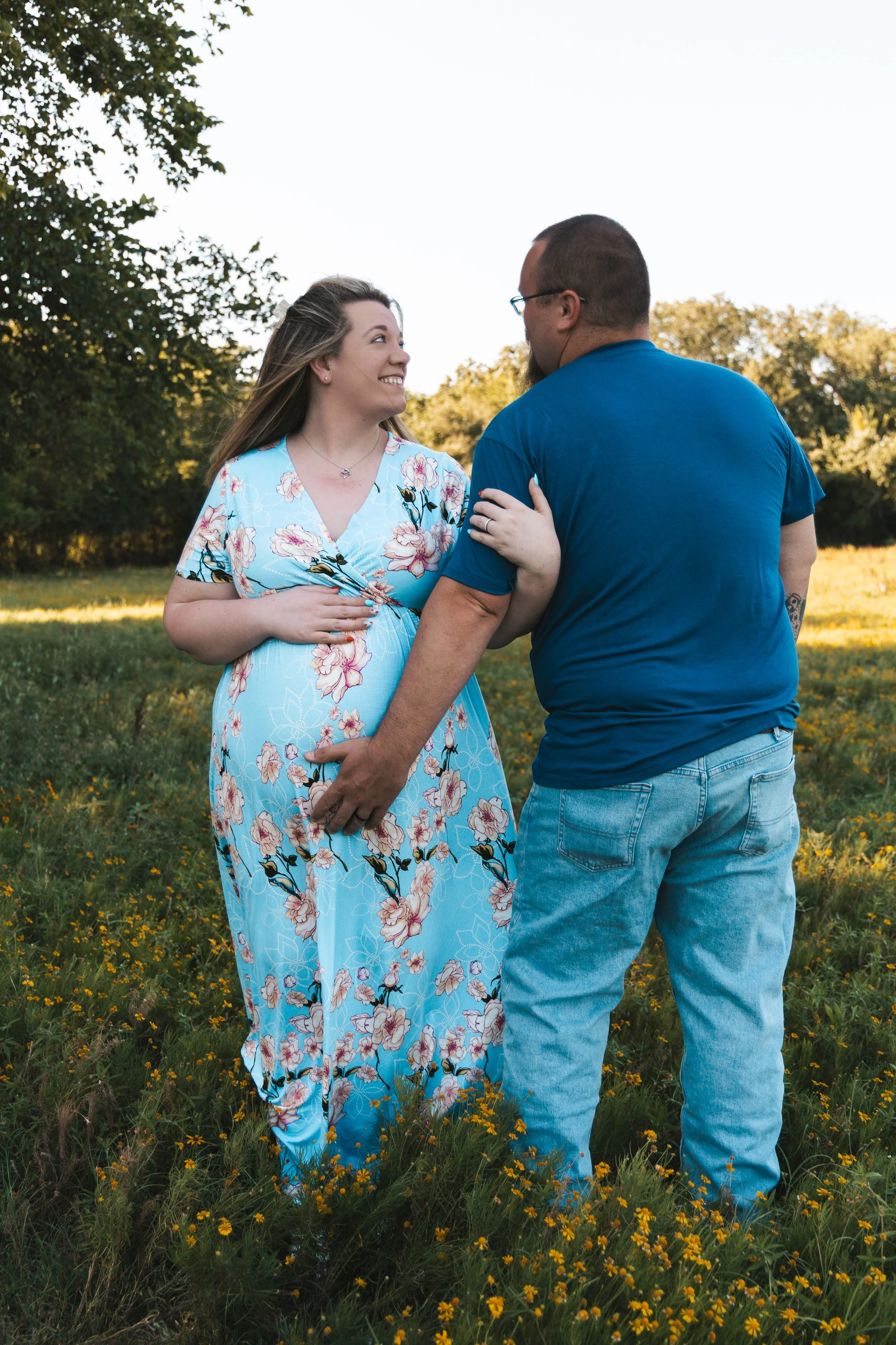 A couple smiling and looking at each other outdoors in a grassy field during daytime, with trees in the background. The woman wears a light blue floral dress, and the man wears a blue t-shirt and jeans.