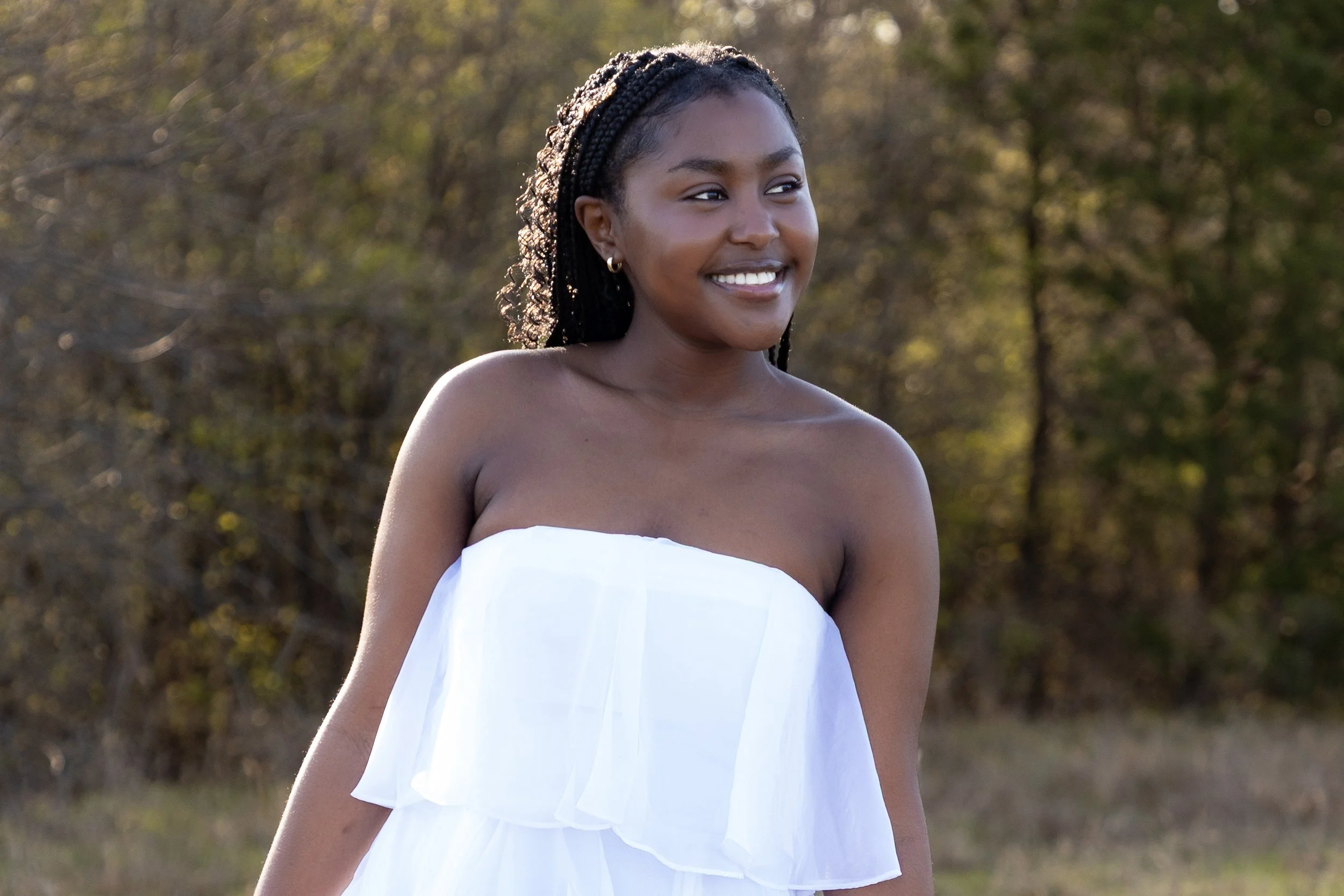A young woman with braided hair wearing a white strapless dress, standing outdoors, smiling, with trees and sunlight in the background.