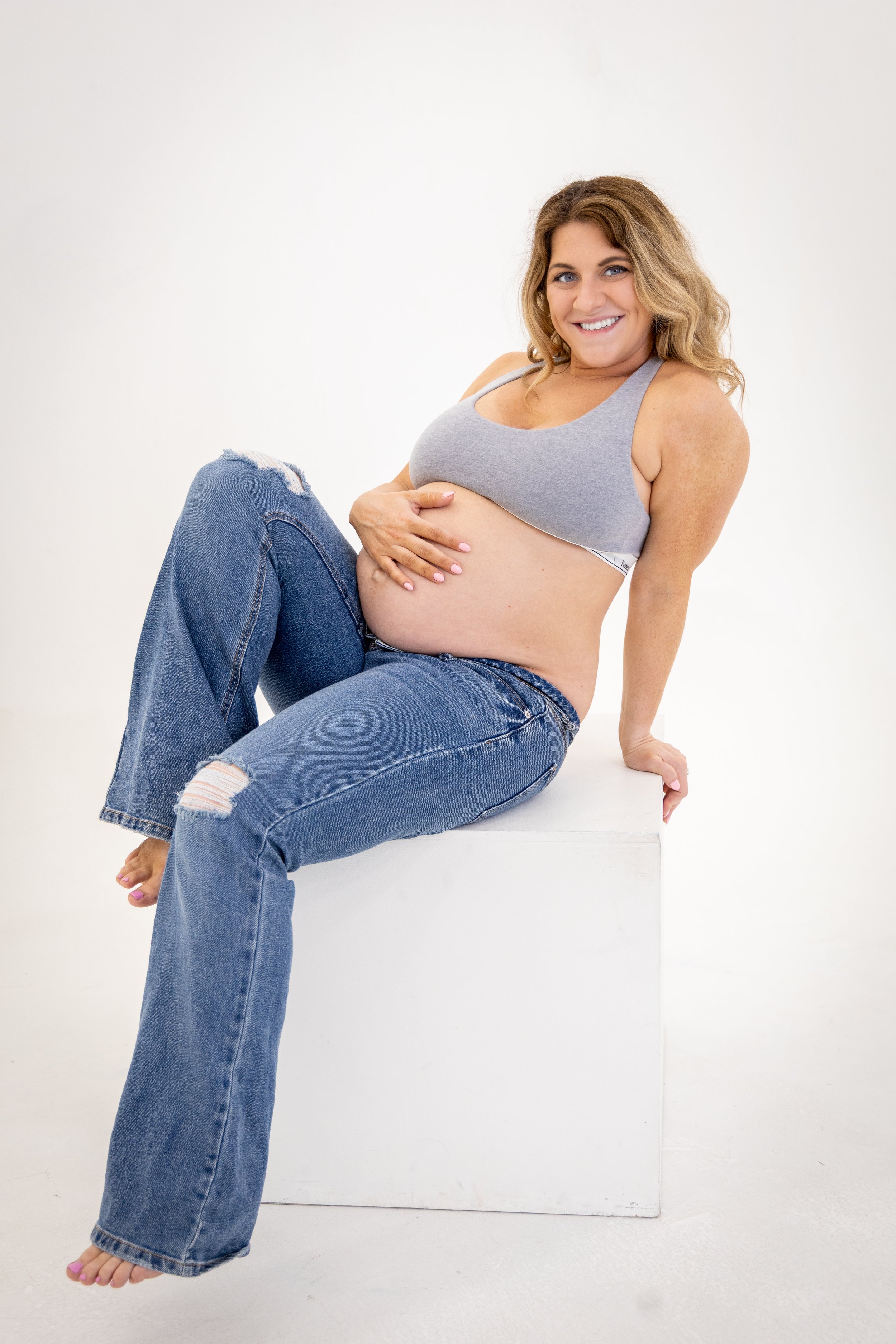 A pregnant woman smiling and sitting on a white box, wearing a gray sports bra and ripped blue jeans, with her right hand on her belly, against a plain white background.