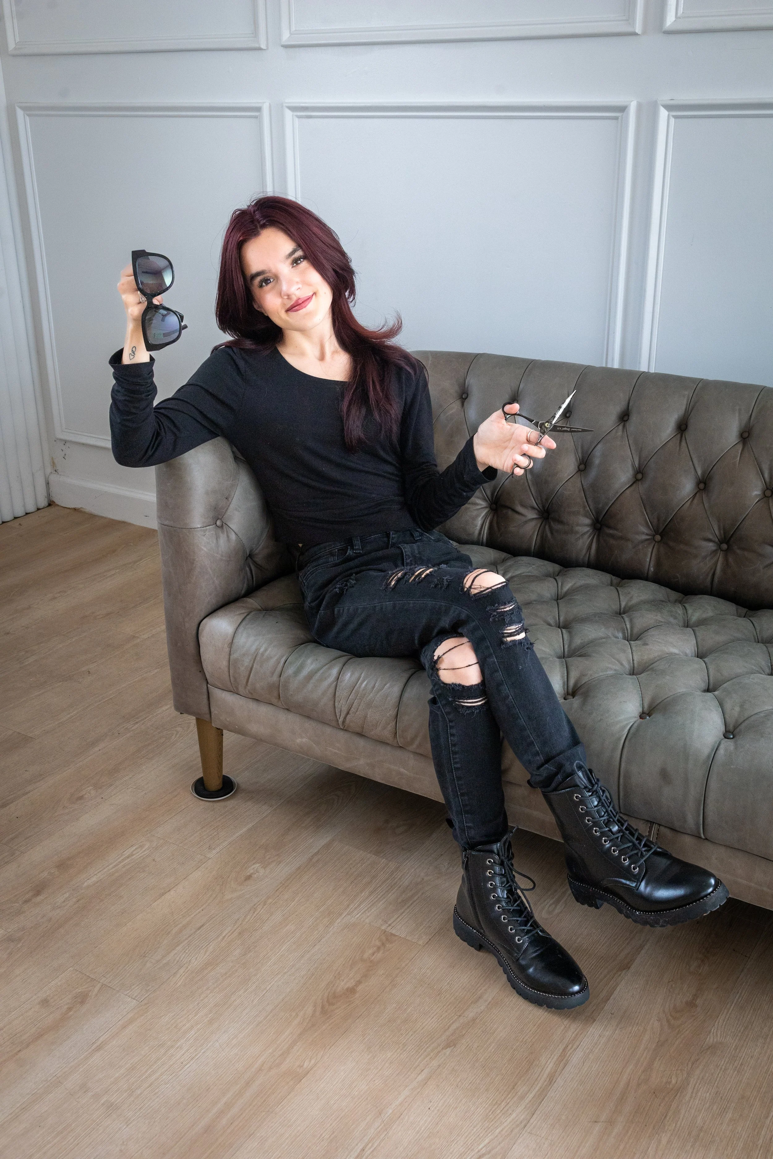 A young woman with red hair sitting on a tan leather couch in a room with white walls. She is wearing a black long-sleeve shirt, ripped black jeans, and black combat boots. She is holding sunglasses in her right hand and scissors in her left hand, sm