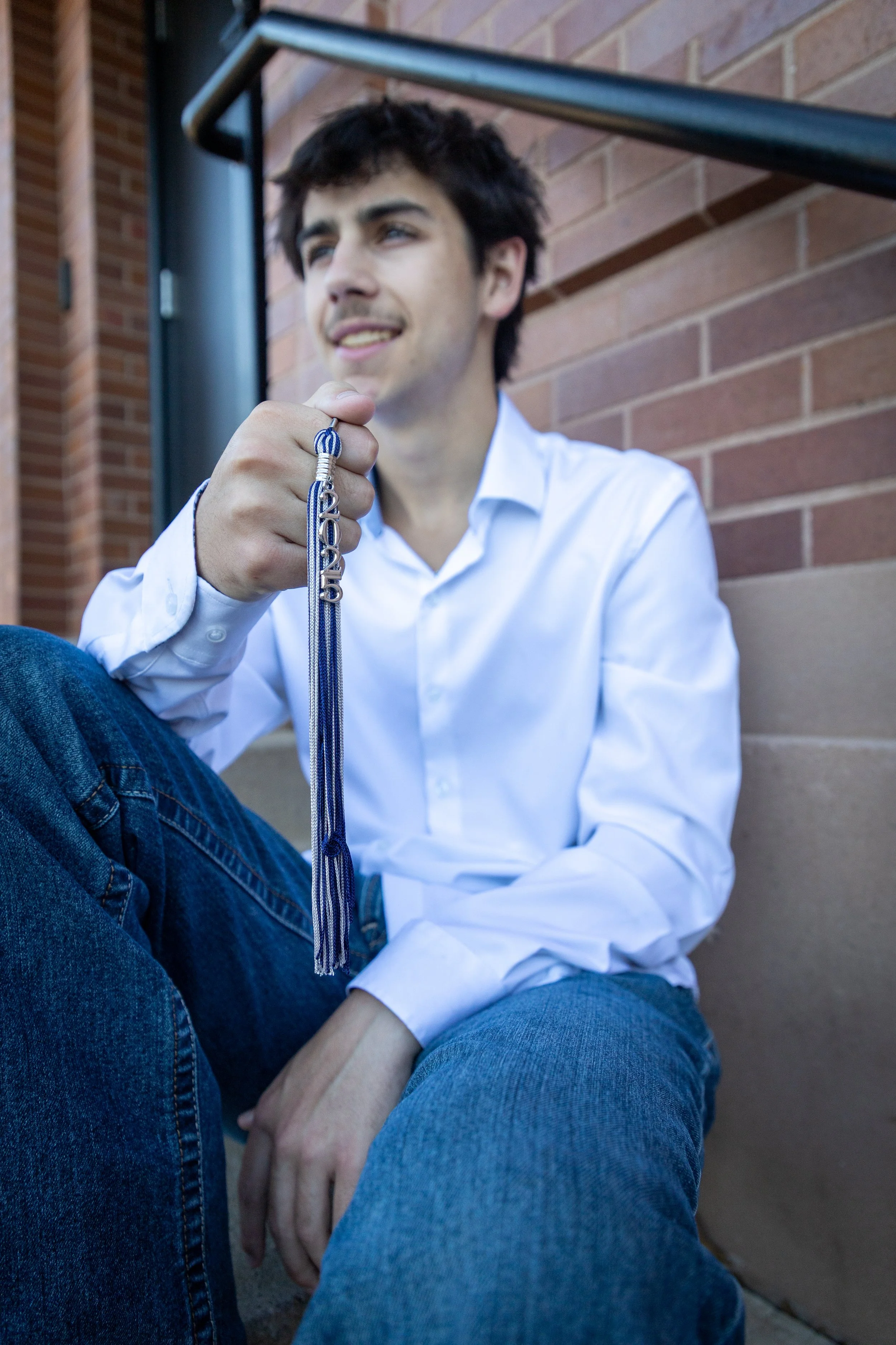 A young man sitting on steps outside a building, holding a graduation tassel, dressed in a white shirt and jeans.