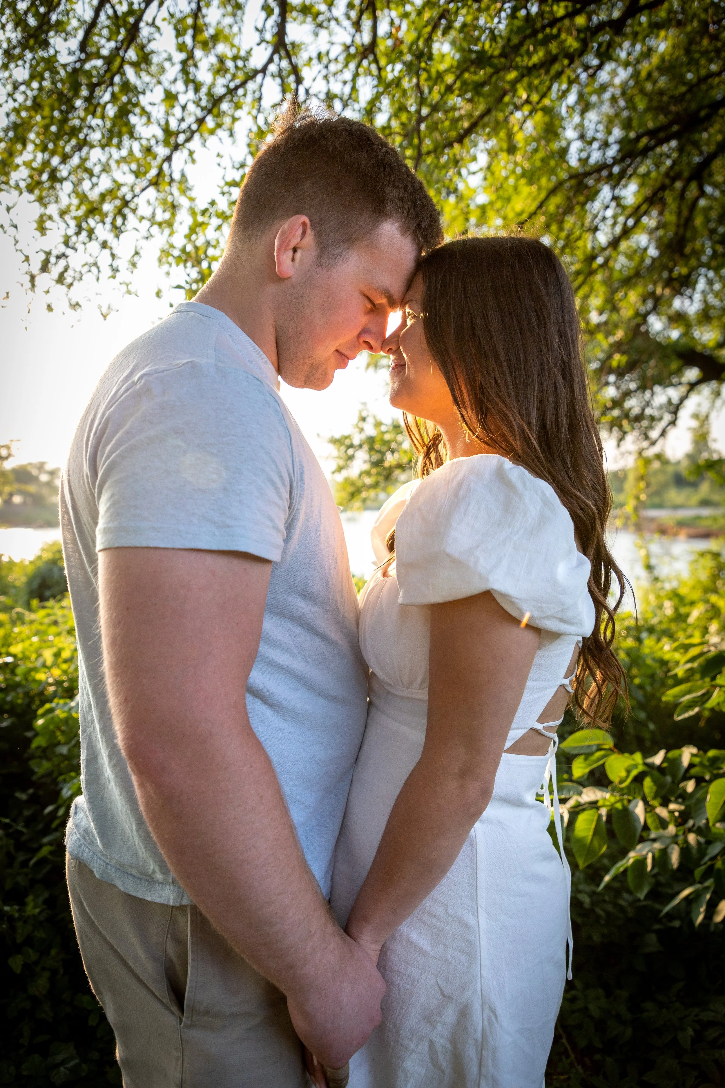 A romantic couple standing outdoors with trees and a river in the background, touching foreheads and holding hands while smiling at each other during sunset.