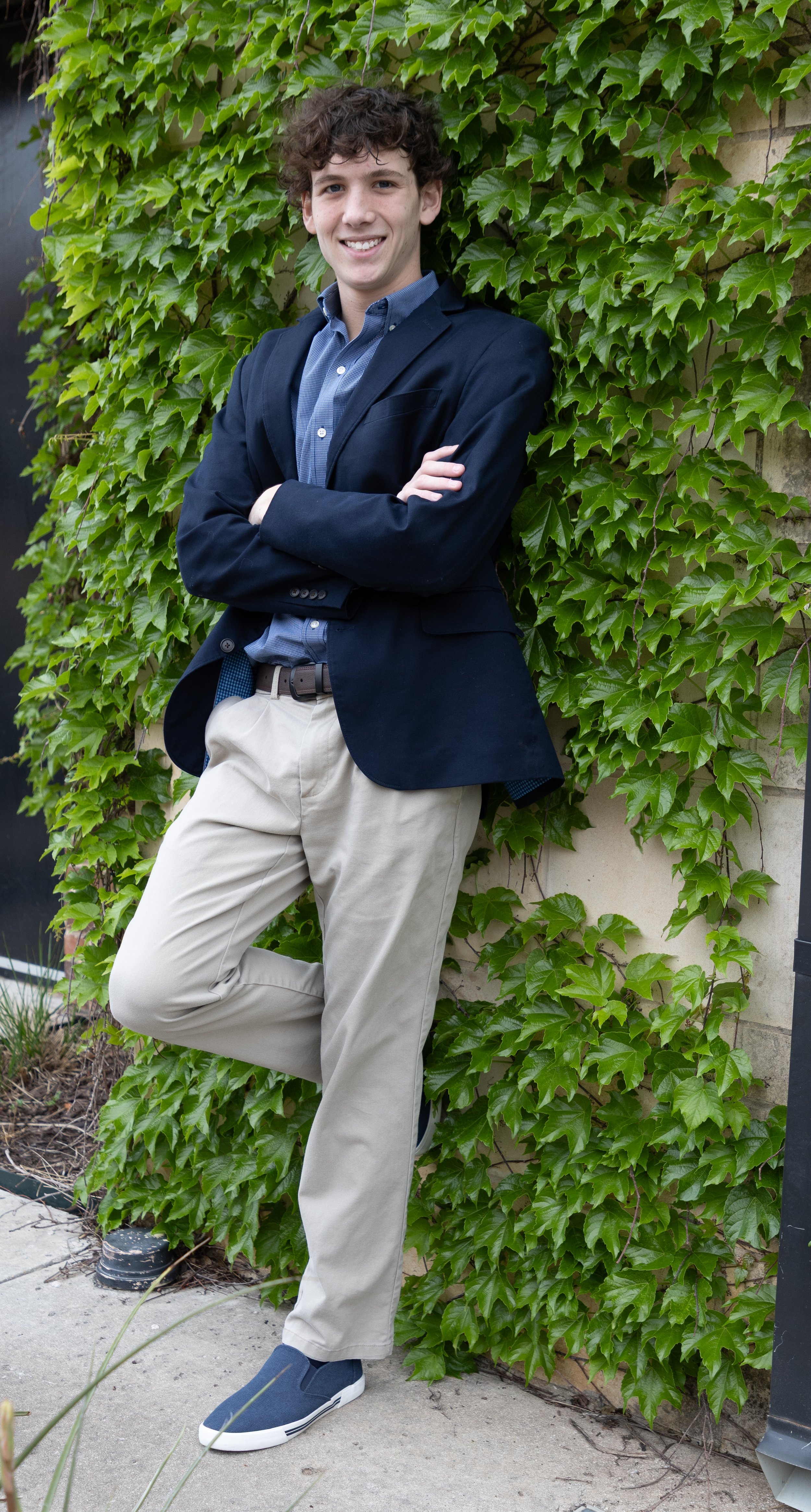 Young man in a navy blazer, blue shirt, and khaki pants leaning against a brick wall covered in green ivy, smiling with arms crossed.