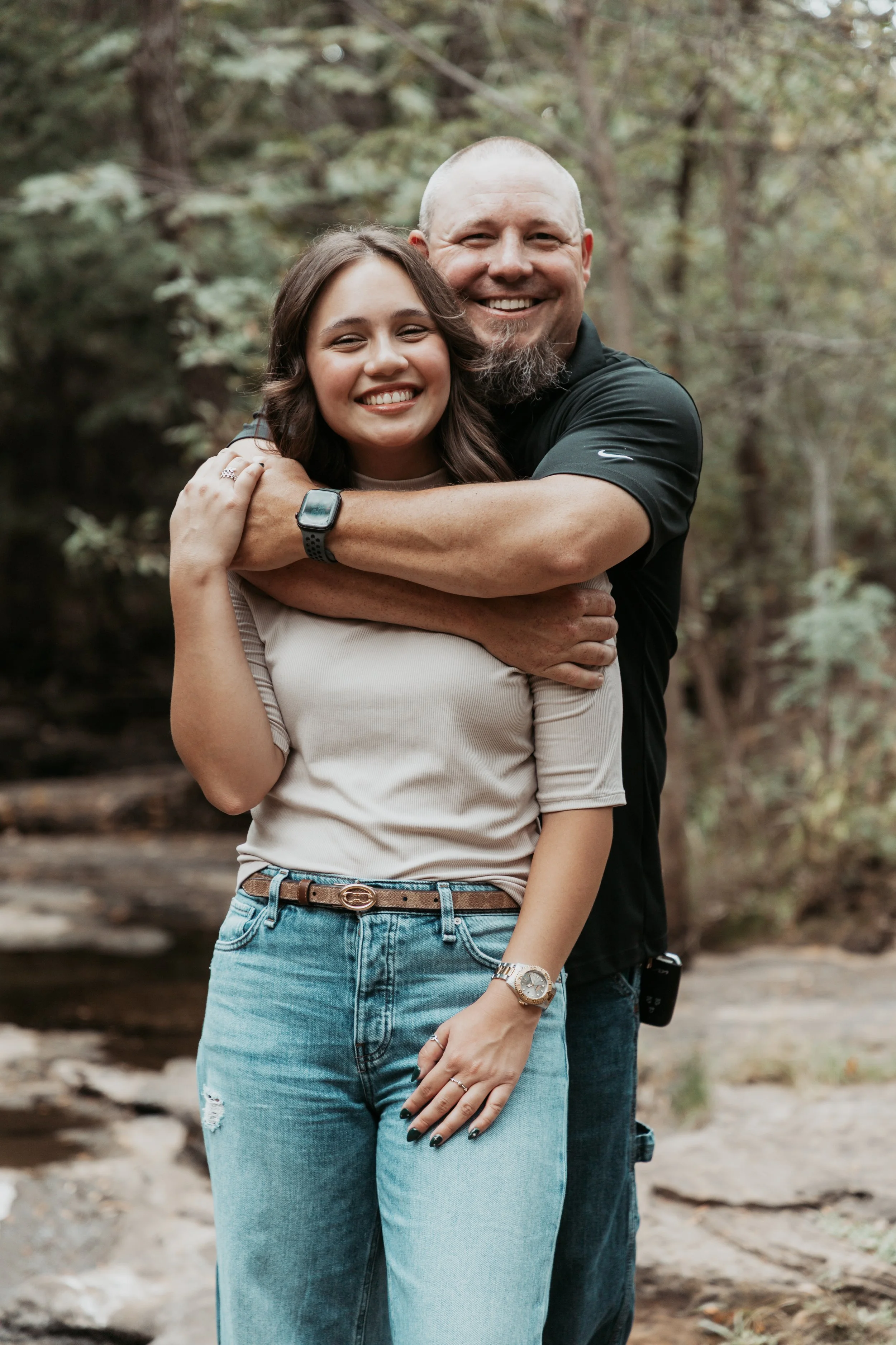 A man and a woman hugging outdoors in a wooded area, both smiling happily.