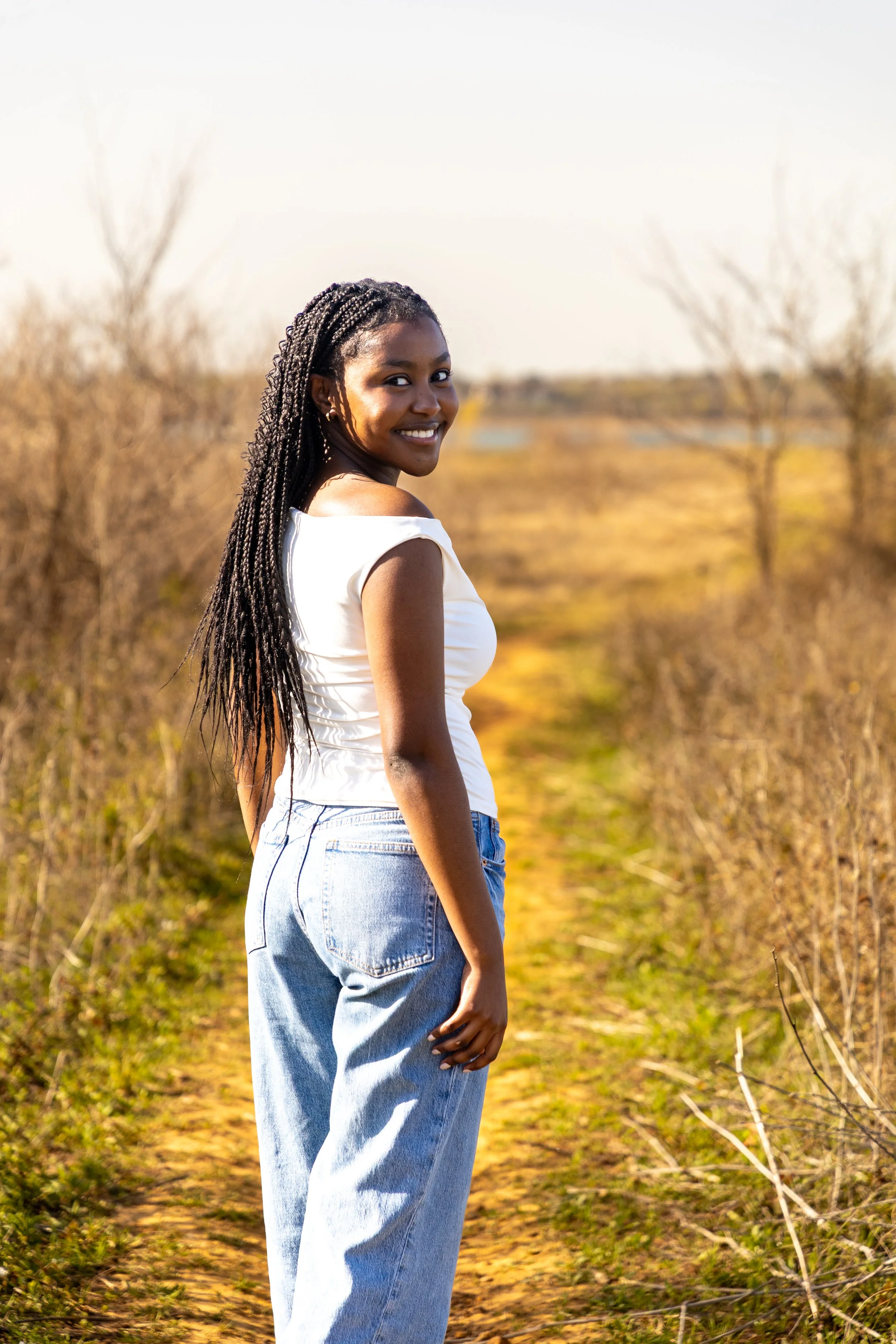 Young woman with long braided hair smiling and standing on a dirt path in a rural area with dry, brown bushes and trees in the background.