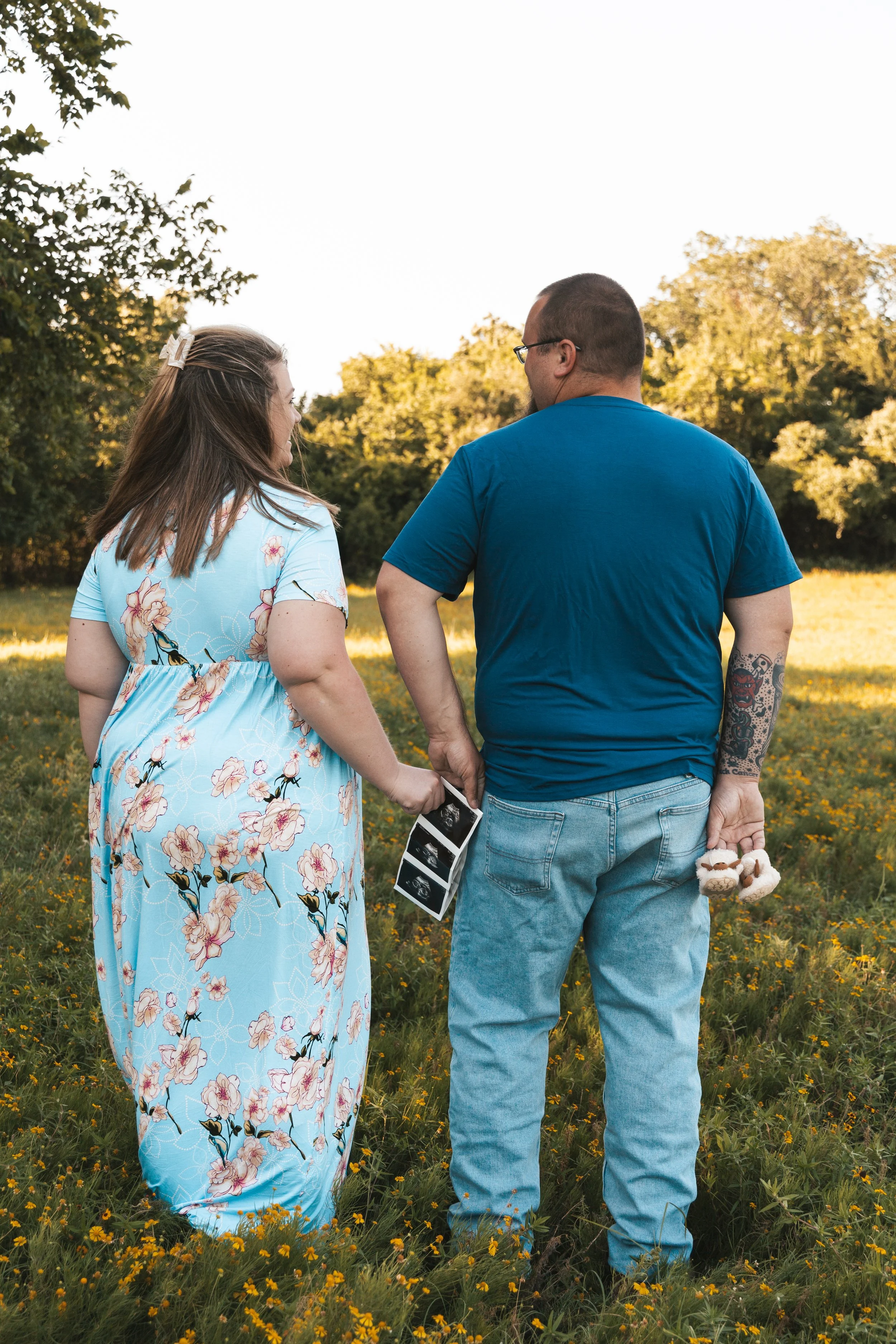A couple holding hands in a field of yellow wildflowers, walking away from the camera. The woman wears a light blue floral dress and the man is in a blue t-shirt and jeans, holding a small stuffed animal and a phone wrapped in a black and white strip