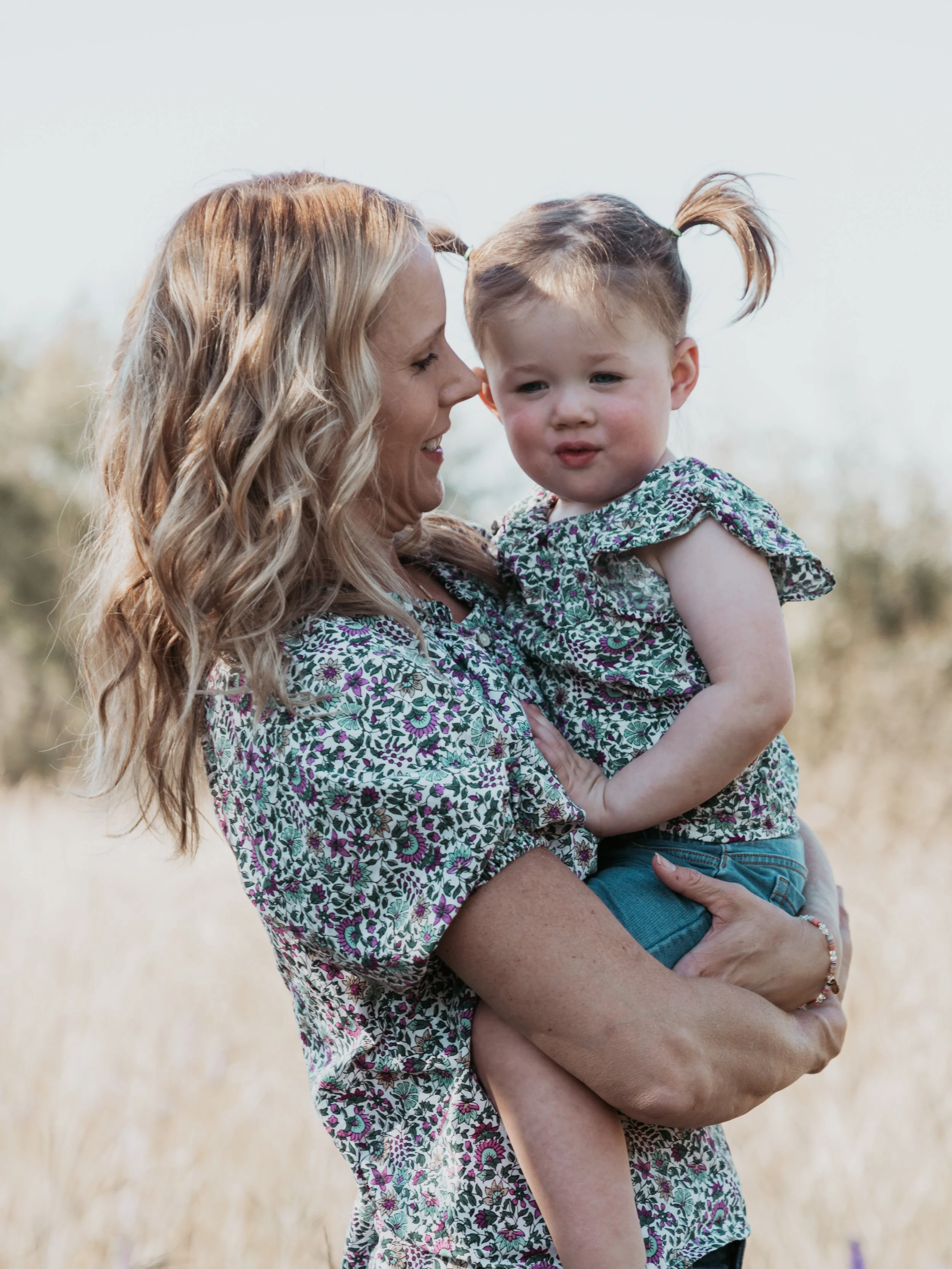 A woman holding a young girl outdoors in a natural setting, both wearing floral patterned tops.