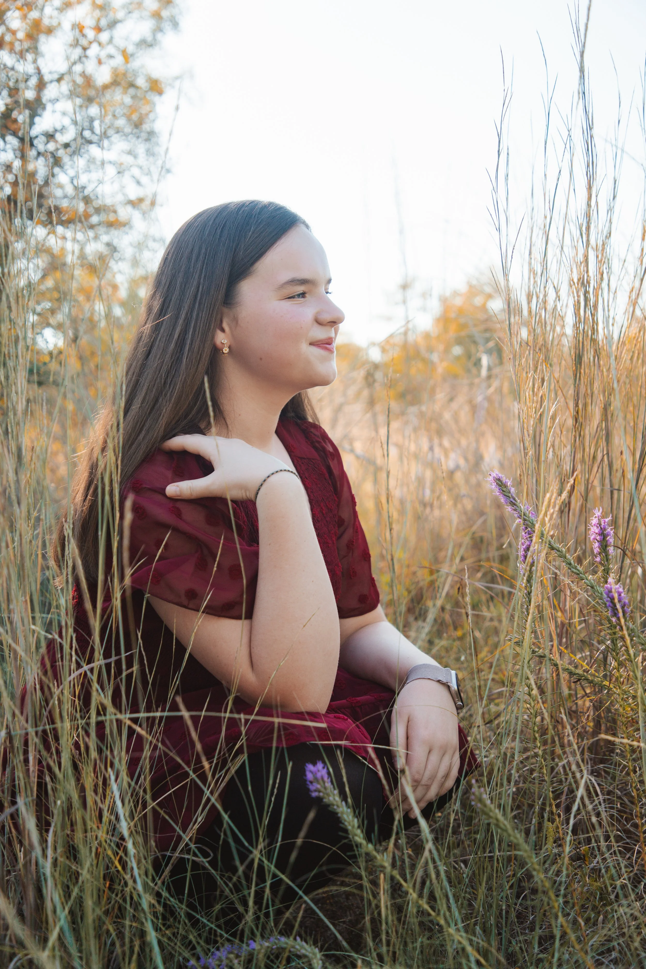 A young woman with long brown hair, wearing a maroon dress, is sitting amidst tall grass and wildflowers in a natural outdoor setting during late afternoon or early evening.