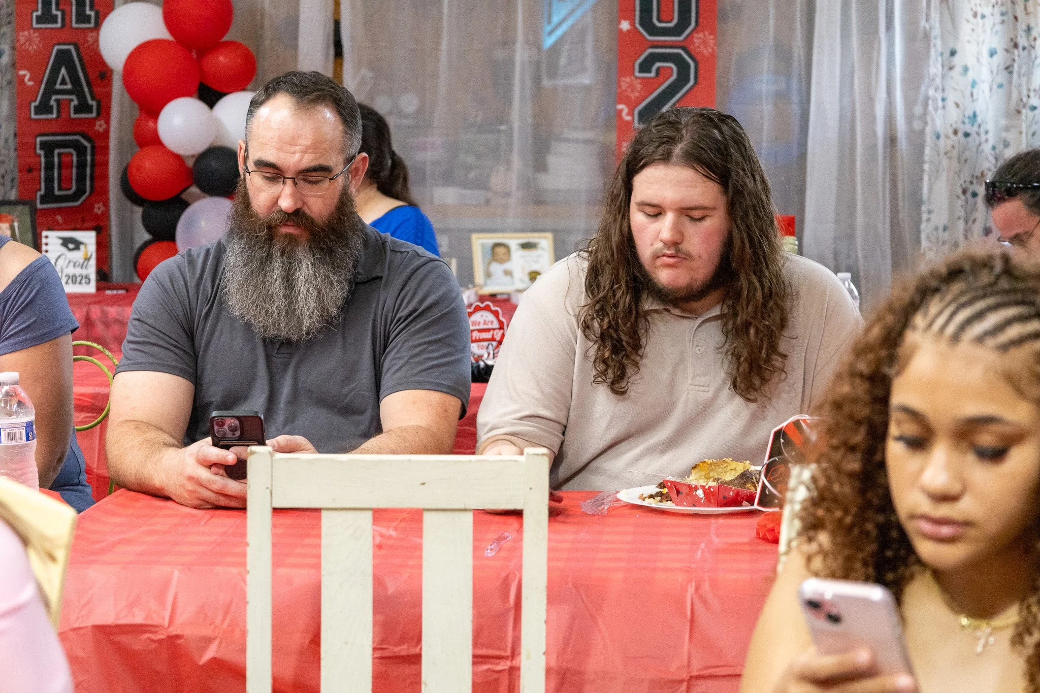 Three people sitting at a table, each looking at their phones during a gathering decorated with red and black balloons and red banners for a graduation celebration, with cake on the table.