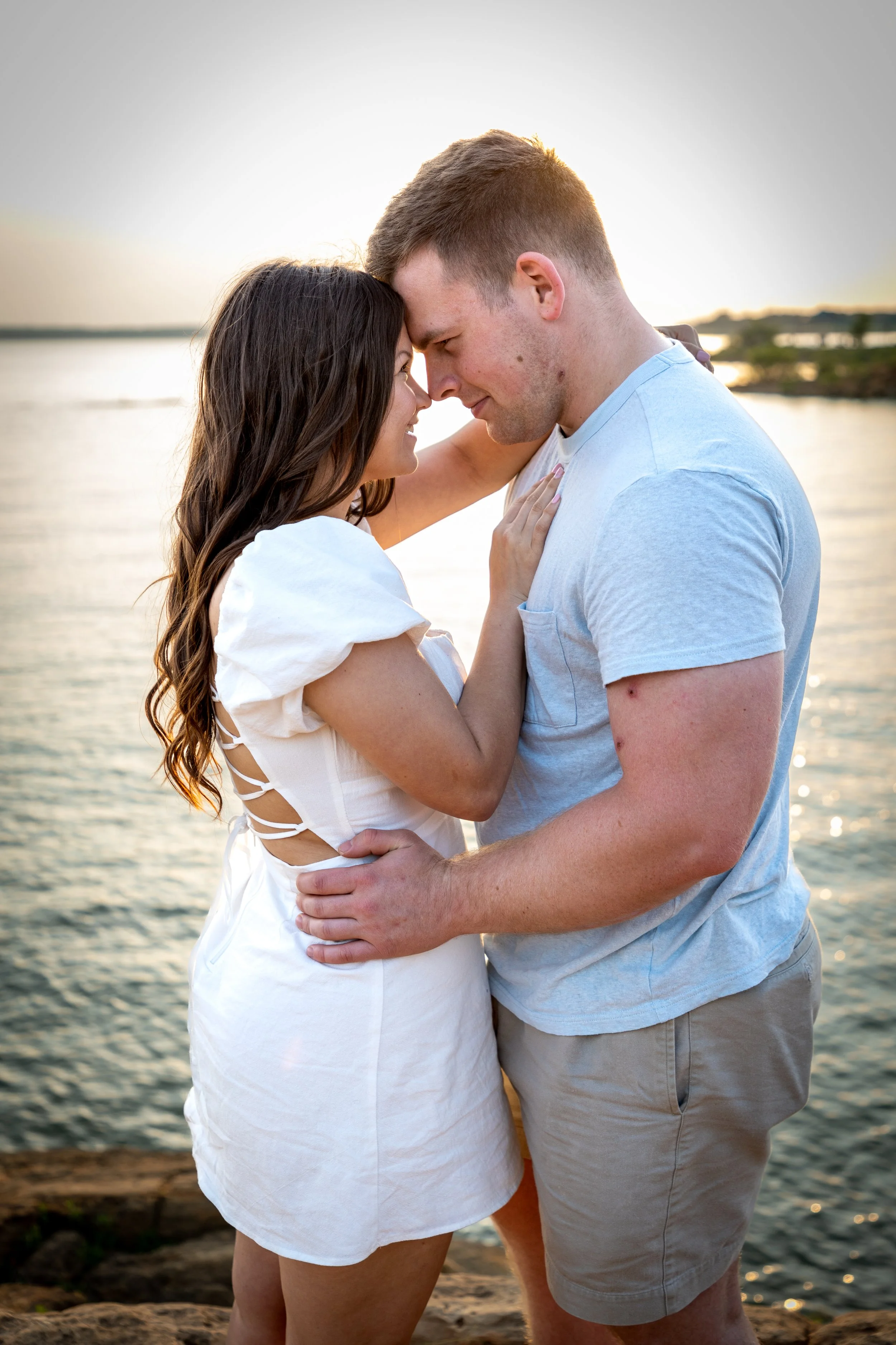 A couple stands close together by the water during sunset, smiling and touching foreheads, with the woman wearing a white dress and the man in a light blue t-shirt and khaki shorts.