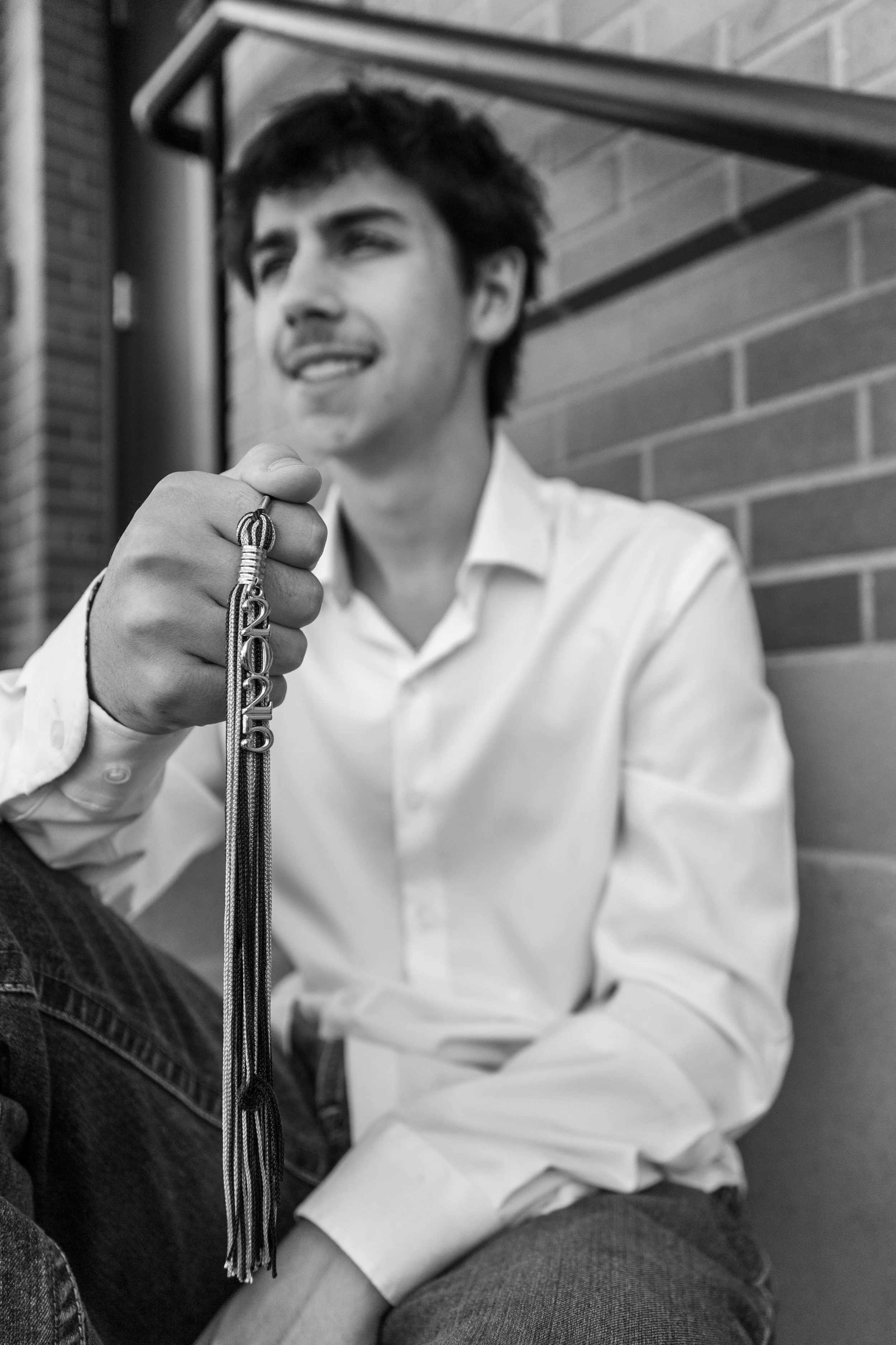 A young man with dark hair, wearing a white shirt, sitting outdoors against a brick wall, holding a necklace with the word "love" on it.