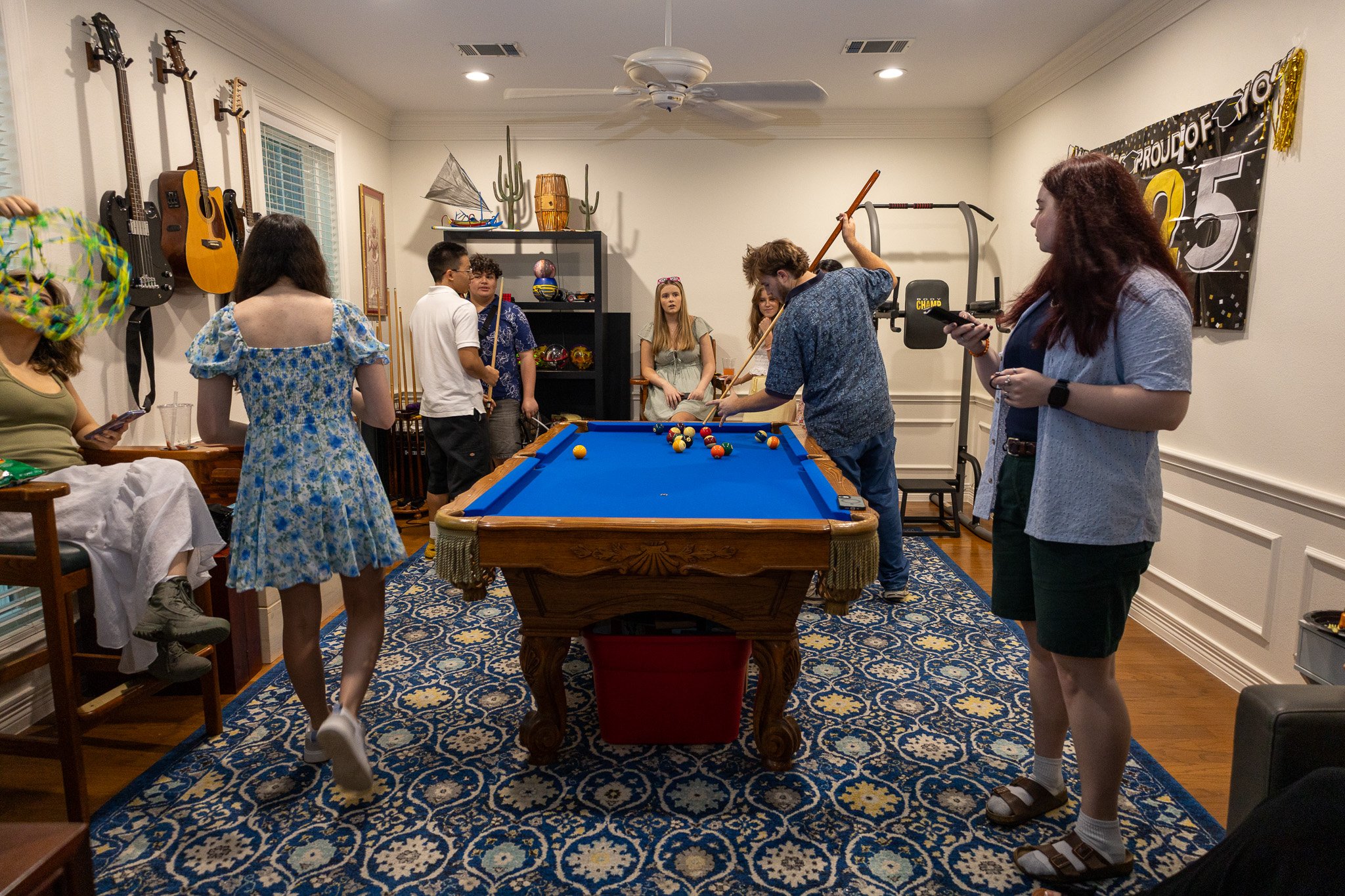 Group of people playing pool in a game room decorated with guitars on the wall, a banner celebrating a 25th birthday, and various decorative items including cactus and boats on a shelf.