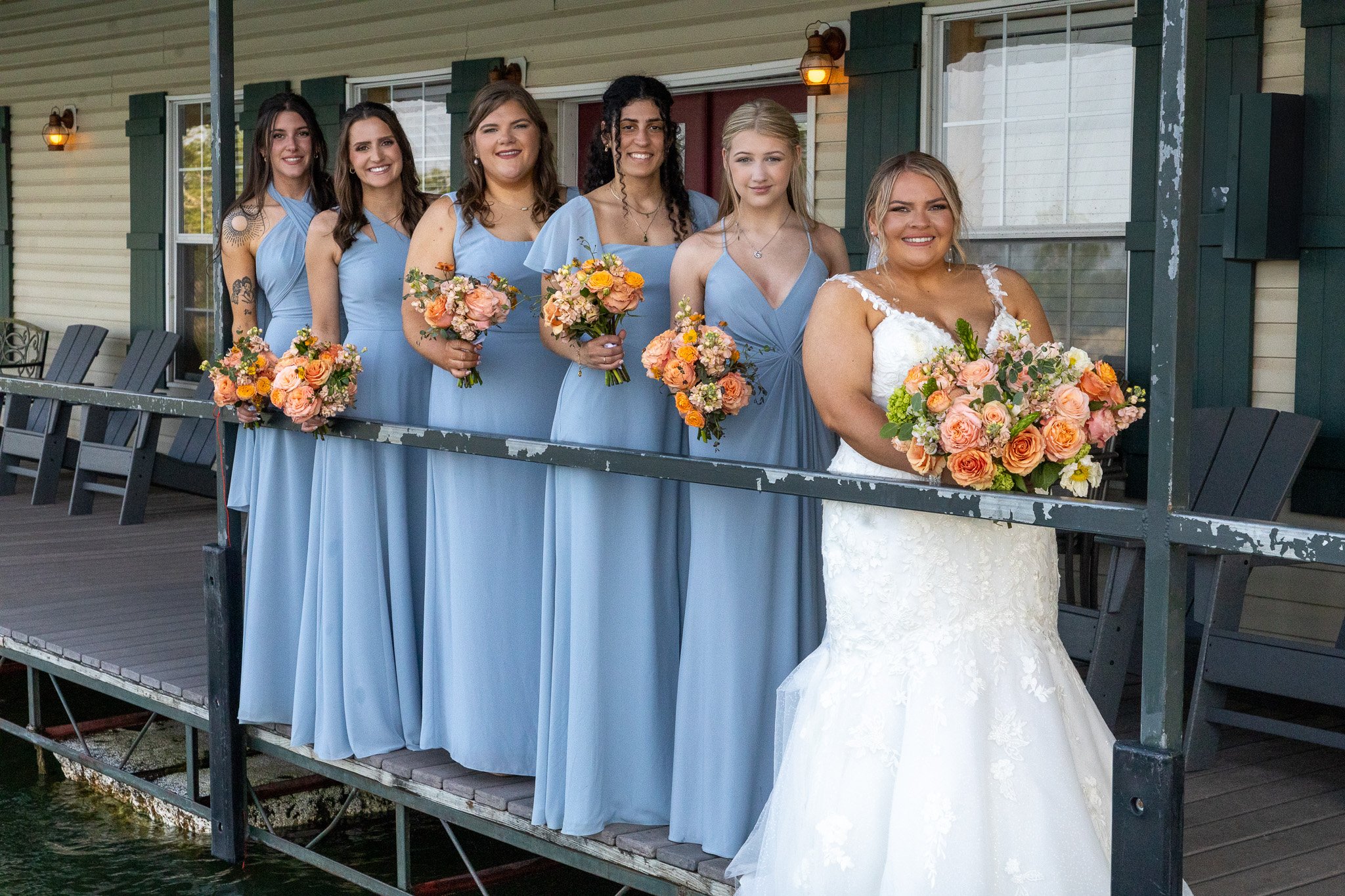 Group of six women, including the bride in a white wedding gown and five bridesmaids in matching light blue dresses, standing on a porch holding bouquets of flowers during a wedding celebration.