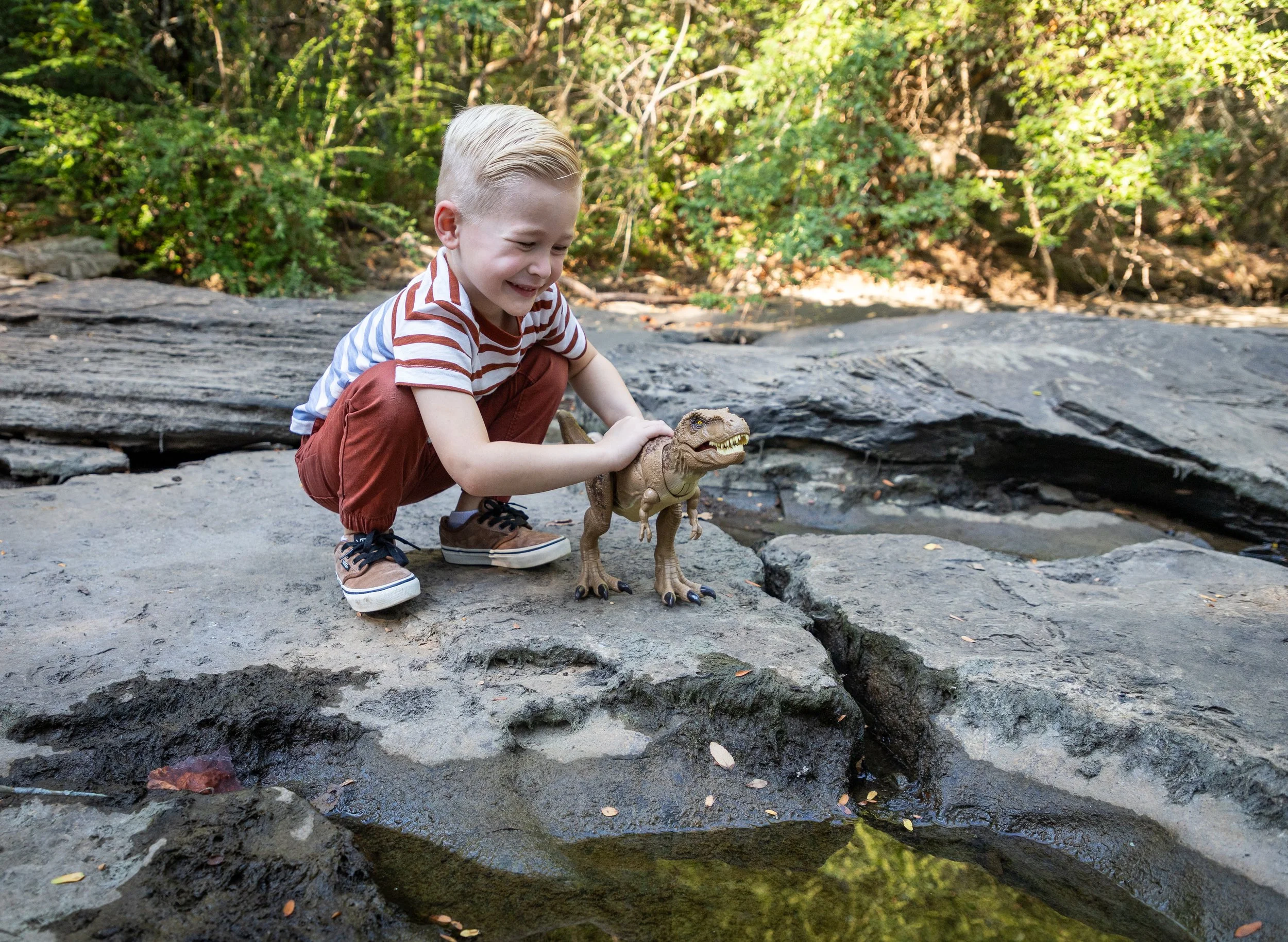 A young boy with blonde hair, wearing a red and white striped shirt and brown pants, crouches on a large rock near a small pond, smiling as he interacts with a toy dinosaur. The background features green trees and foliage.