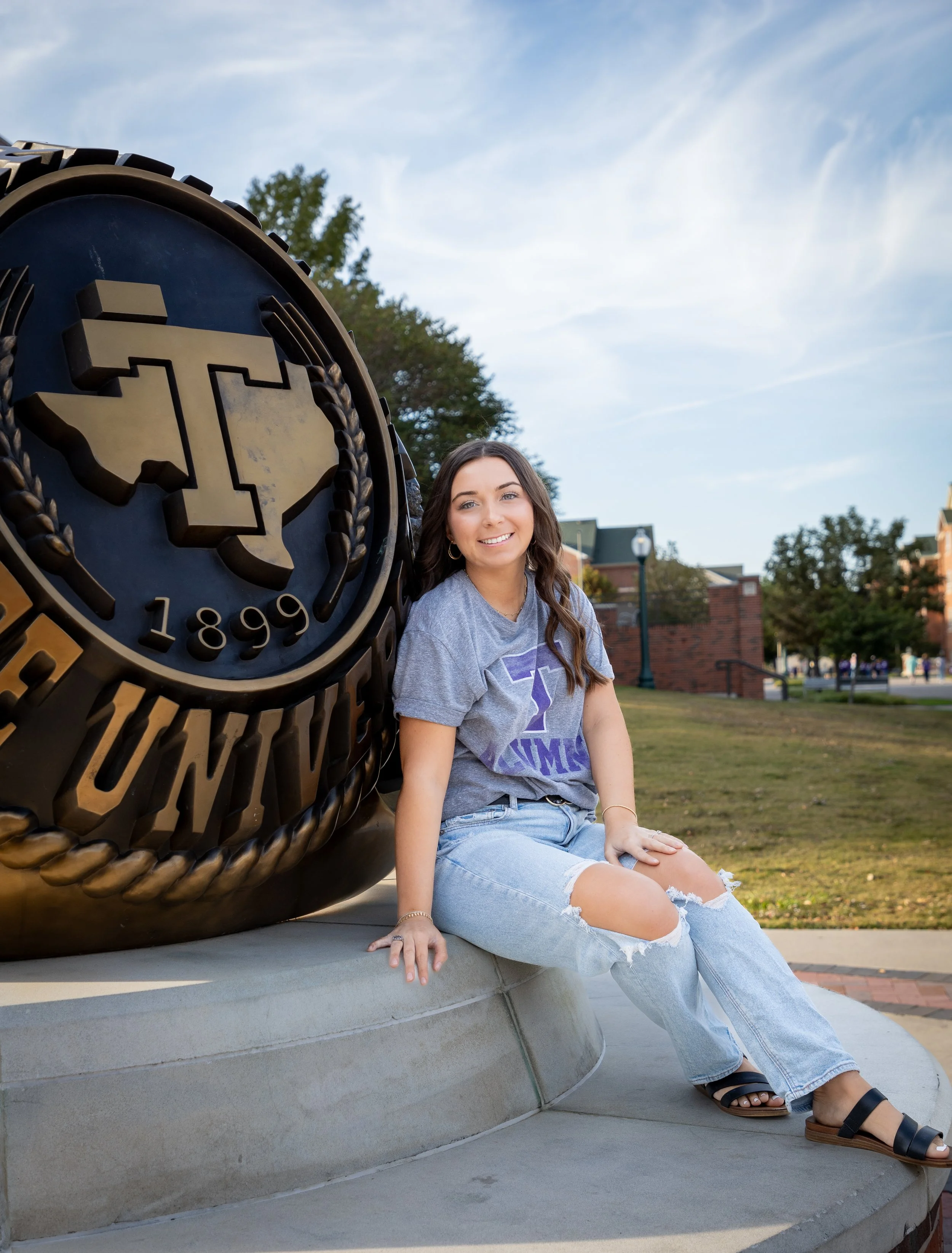 A young woman sitting on a concrete platform, next to a large university emblem sculpture, on a college campus with trees and brick buildings in the background.