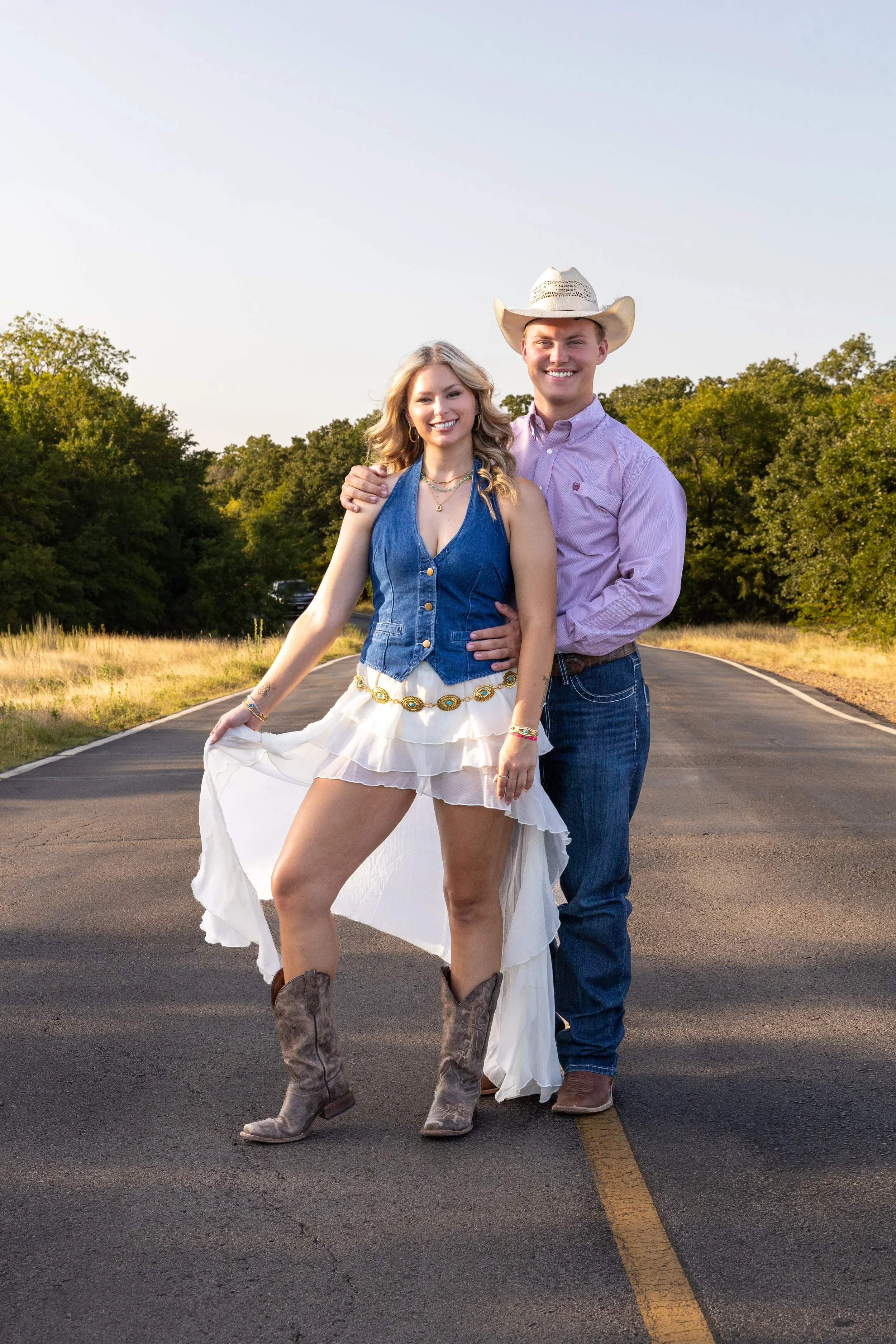 A young couple standing on a rural road, with the woman in cowboy boots and a white dress, and the man wearing a cowboy hat and jeans, both smiling at the camera during sunset.
