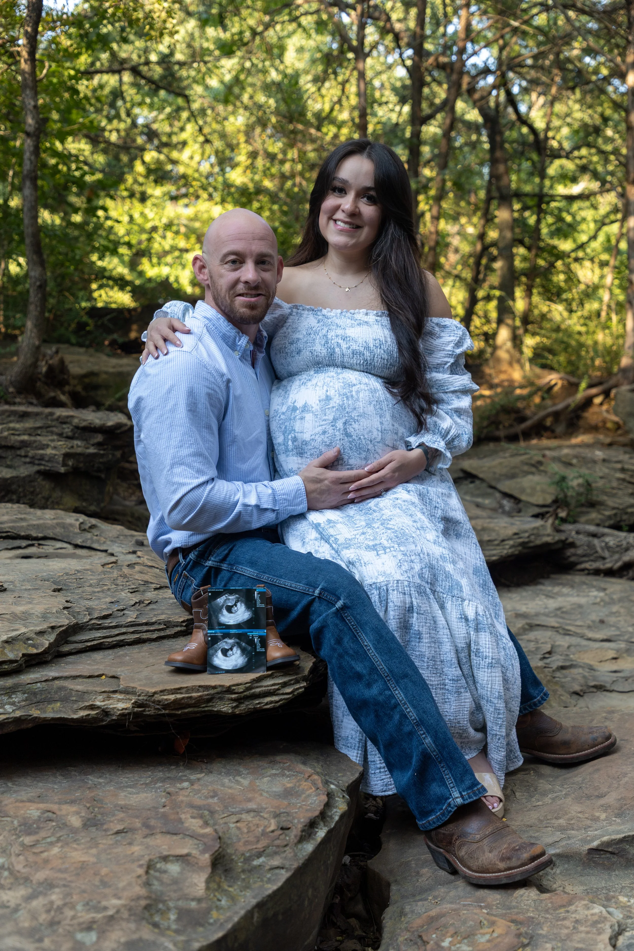 A pregnant woman and a man sitting outdoors on rocks surrounded by trees. The woman is wearing a light off-shoulder dress, and the man is dressed in a light blue shirt and jeans. They are smiling, and the man has his arm around the woman. Two ultraso
