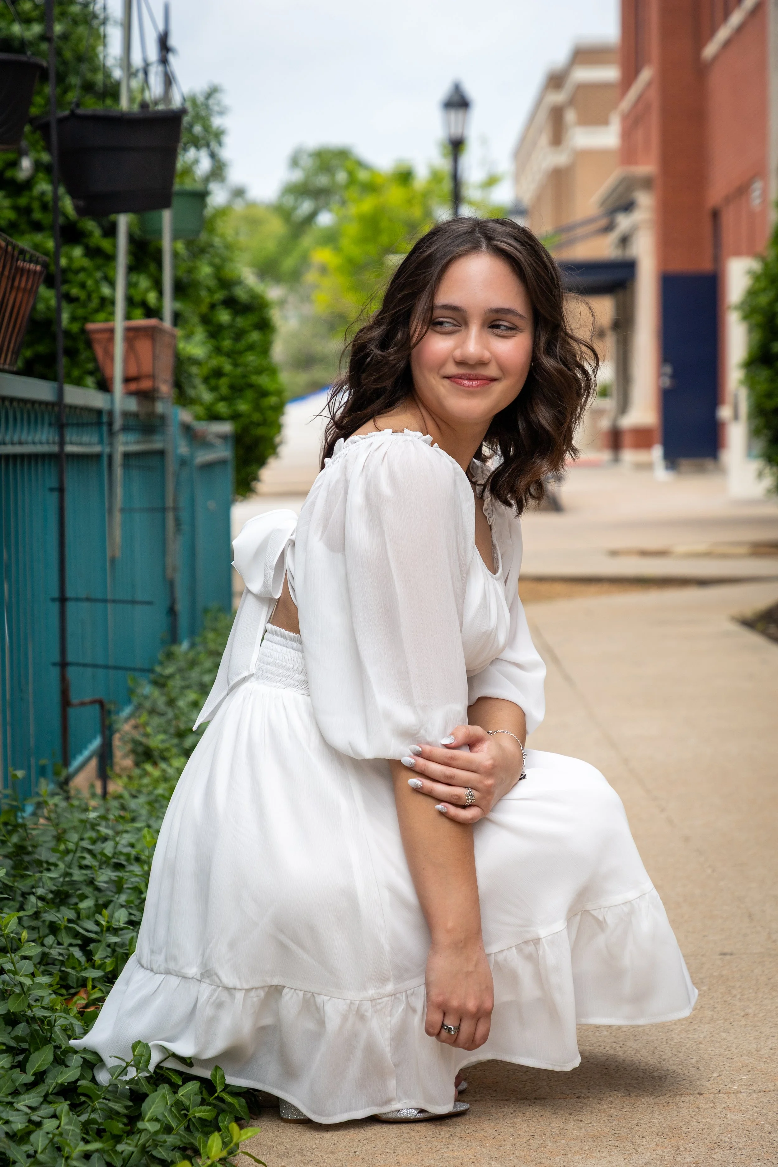 A young woman with curly brown hair, wearing a white dress, sitting on the sidewalk while smiling softly and looking to her right.