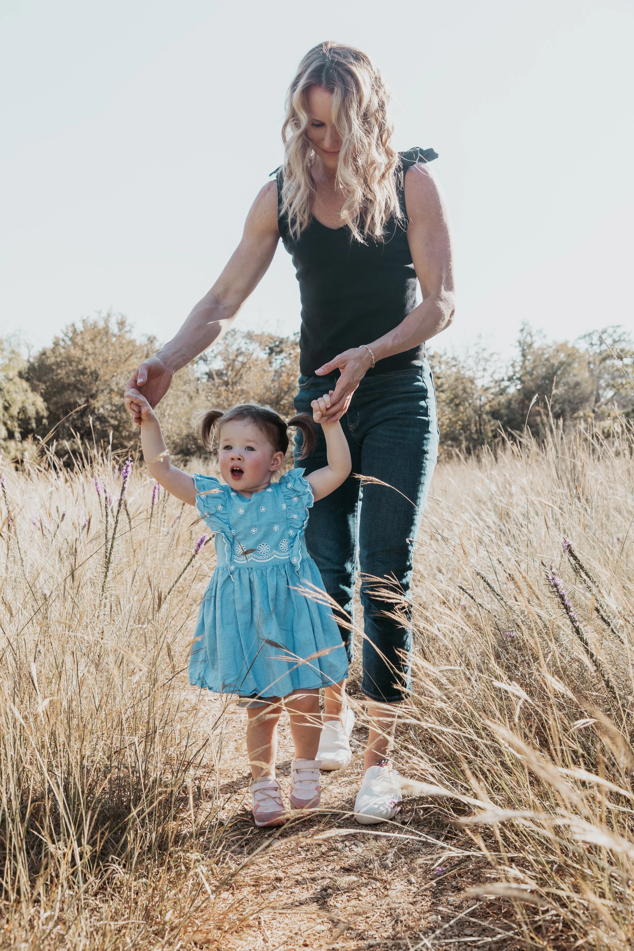 A woman with blonde, wavy hair in a black top and jeans holding hands with a young girl in a blue dress walking through a field of tall grass on a sunny day.