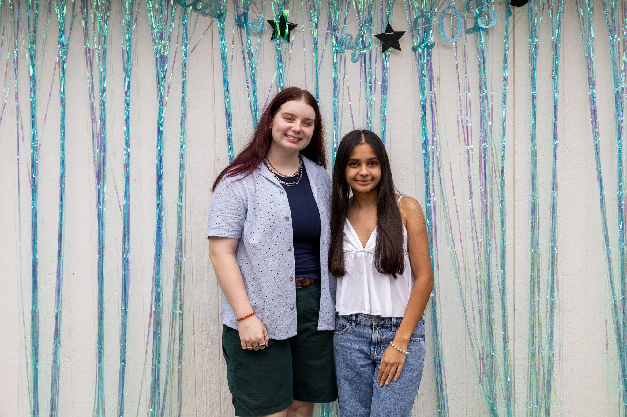 Two young women standing side by side in front of a backdrop decorated with iridescent streamers and black star-shaped balloons, celebrating an event.