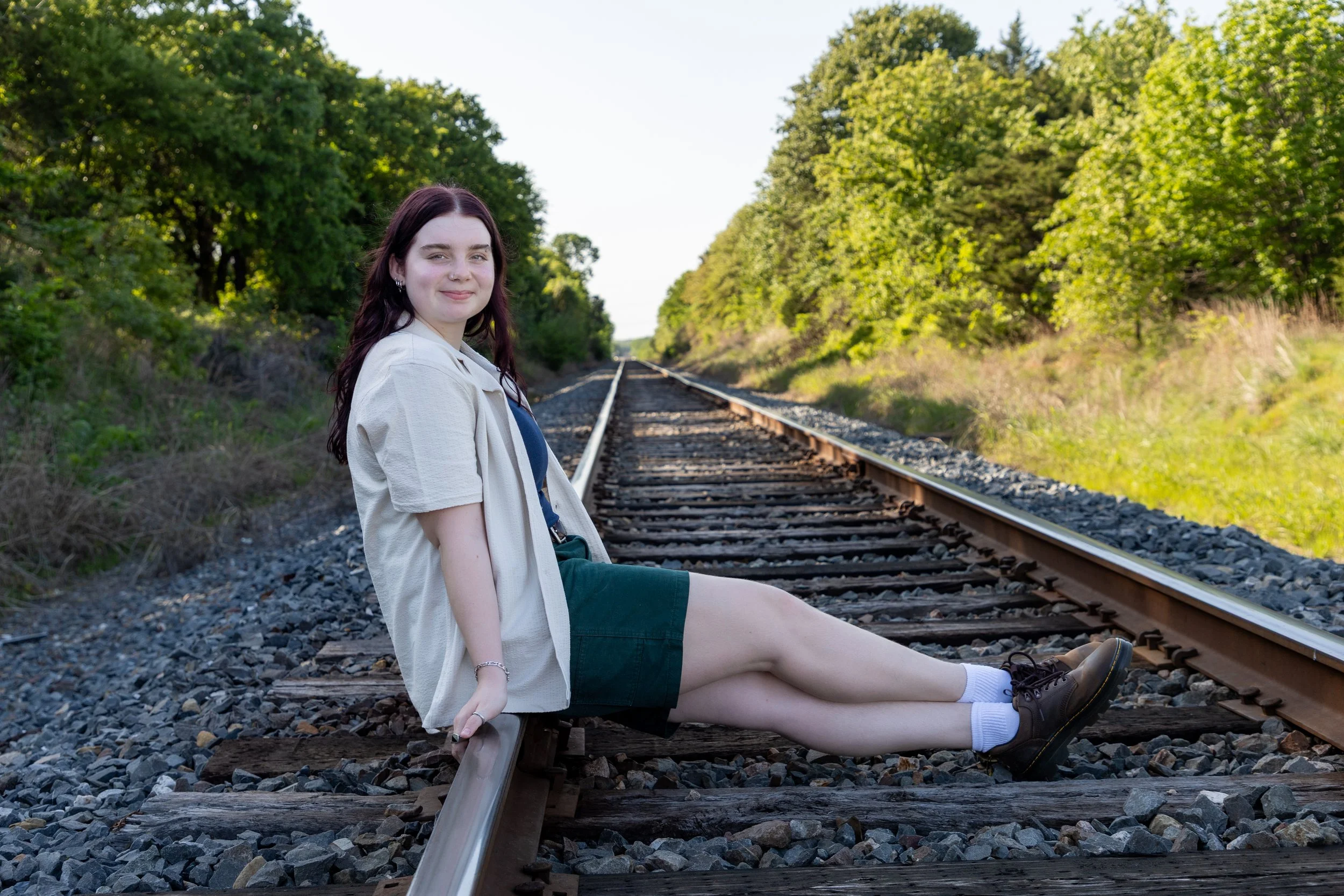 A young woman sitting on railroad tracks surrounded by green trees and grass, smiling at the camera.