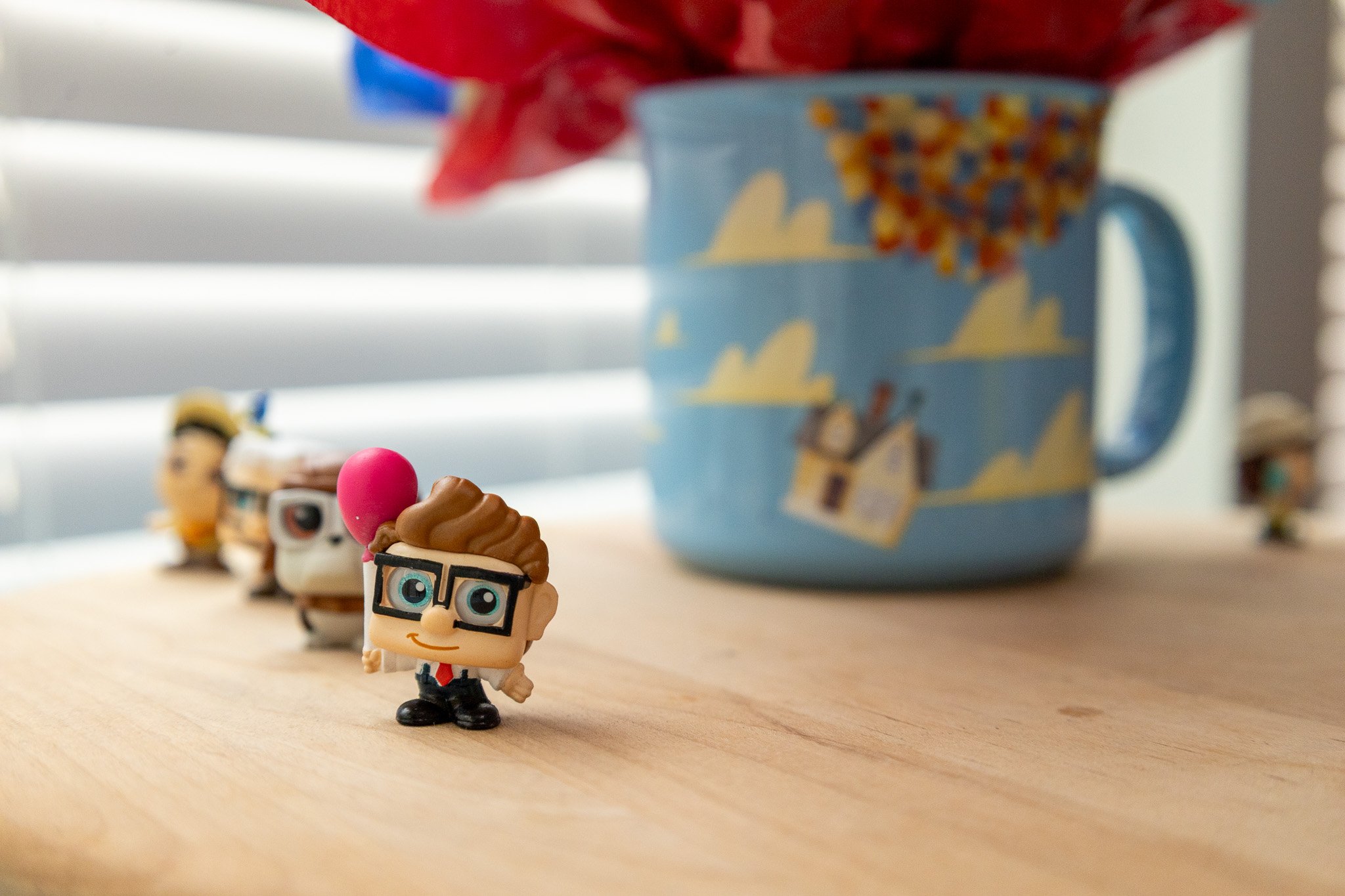A small toy figure with glasses, a suit, and a red tie standing on a wooden surface. Behind it, other similar toy figures are arranged in a line. To the right, there is a blue mug with a cartoon design featuring figures and clouds.