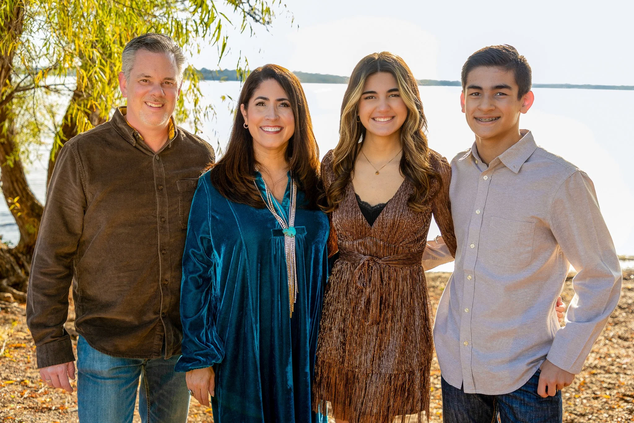 A family of four standing outdoors near a lake with trees in the background, smiling at the camera