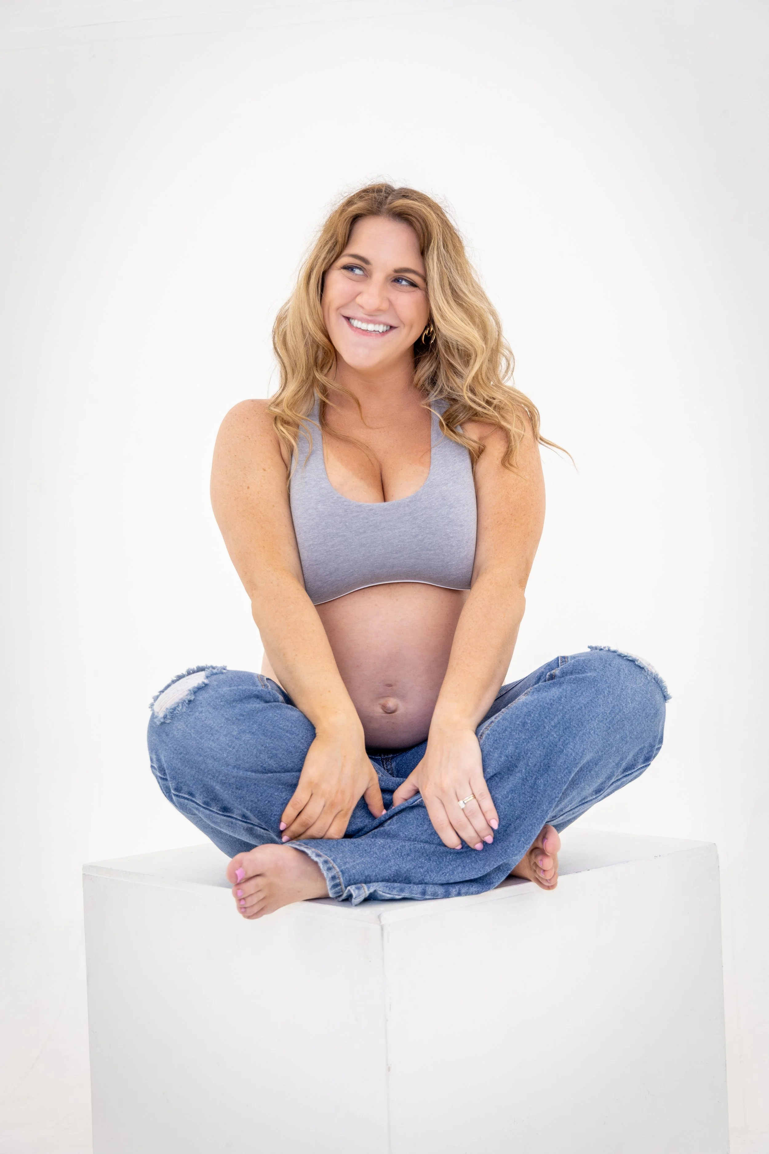 A pregnant woman with wavy blonde hair sitting cross-legged on a white platform, smiling, wearing a gray sleeveless top and ripped jeans, against a plain white background.