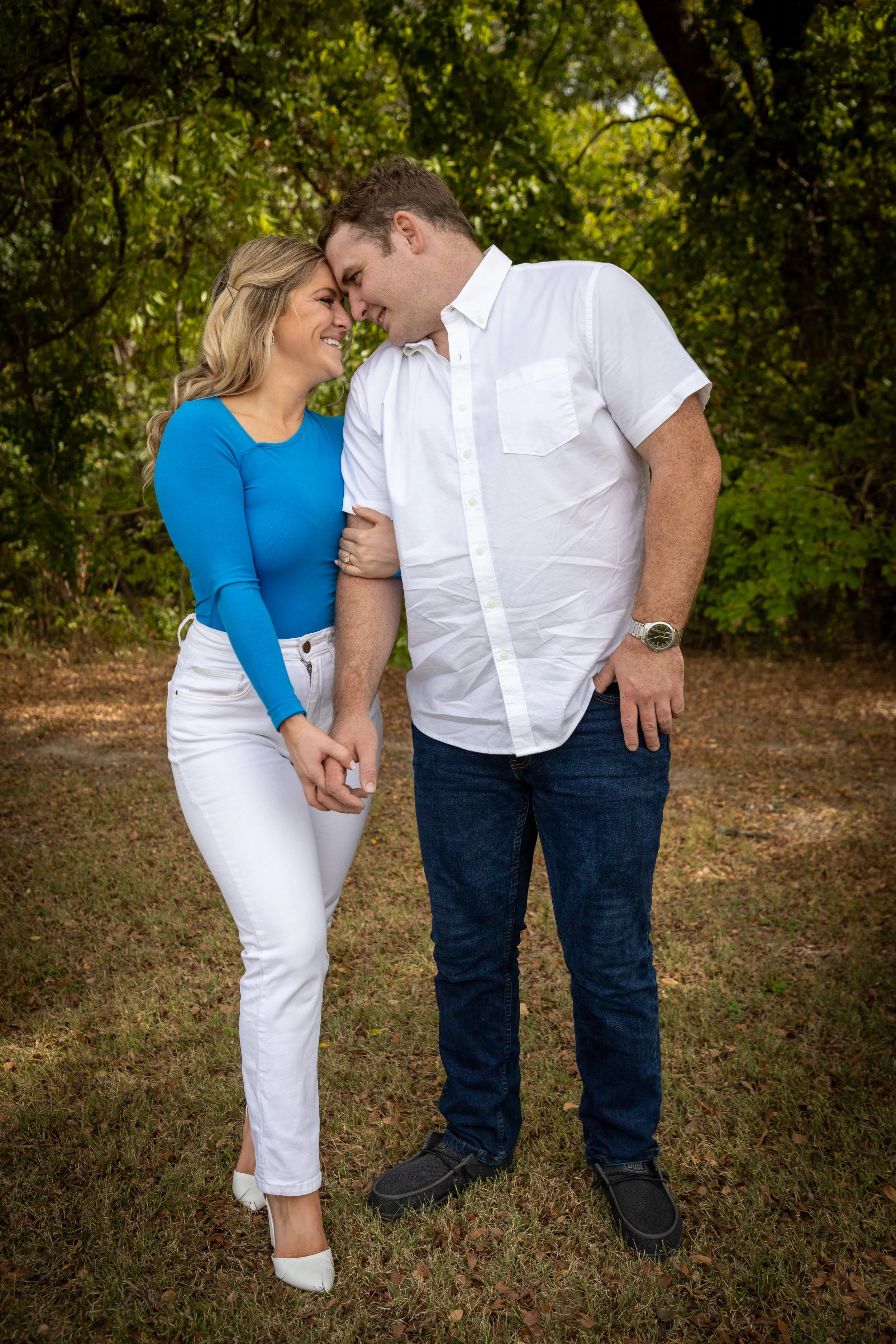 A couple holding hands and leaning their foreheads together outdoors in a wooded area, smiling at each other.
