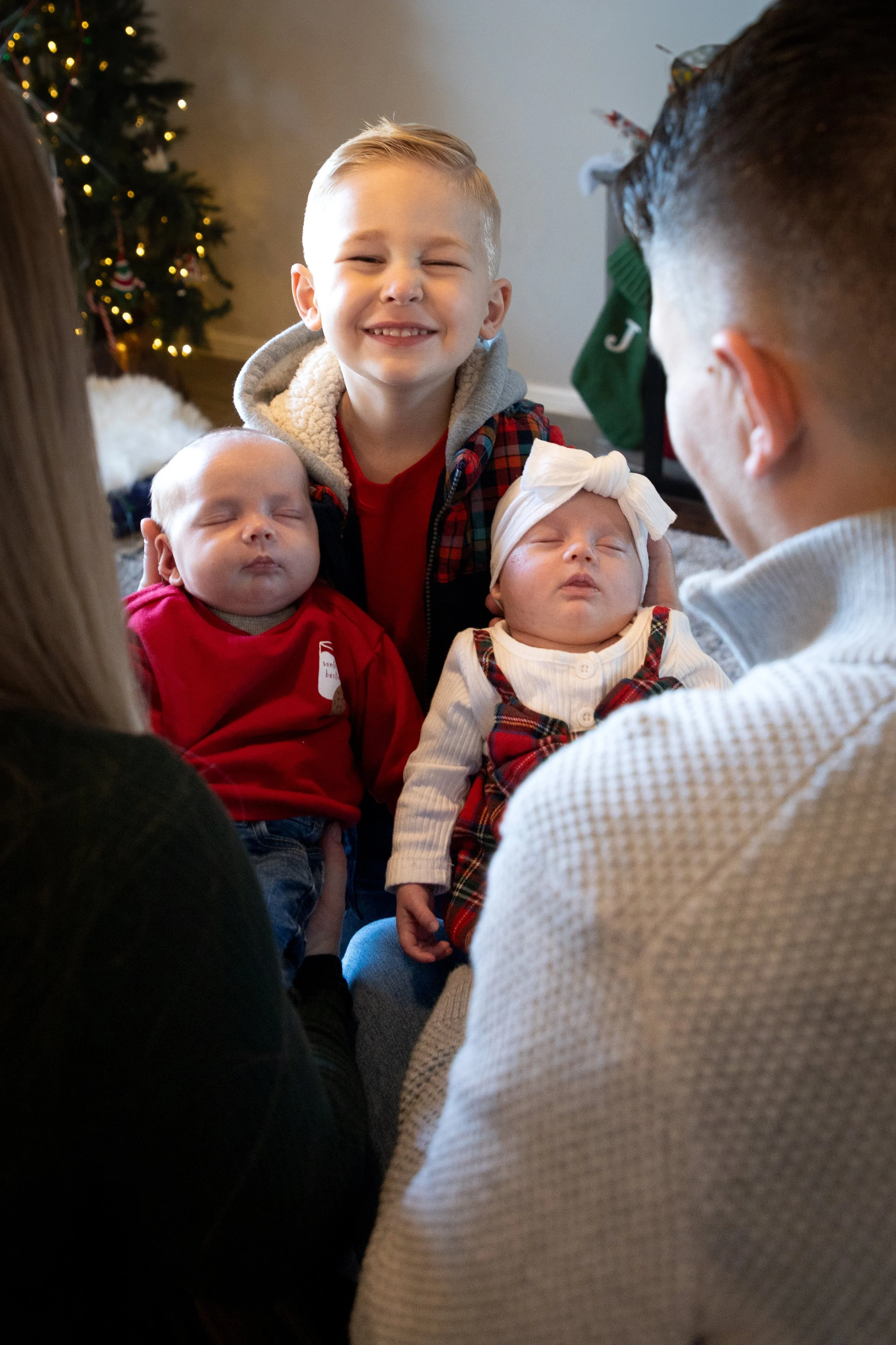 Young boy and family holding two sleeping babies at Christmas with decorated Christmas tree in background.