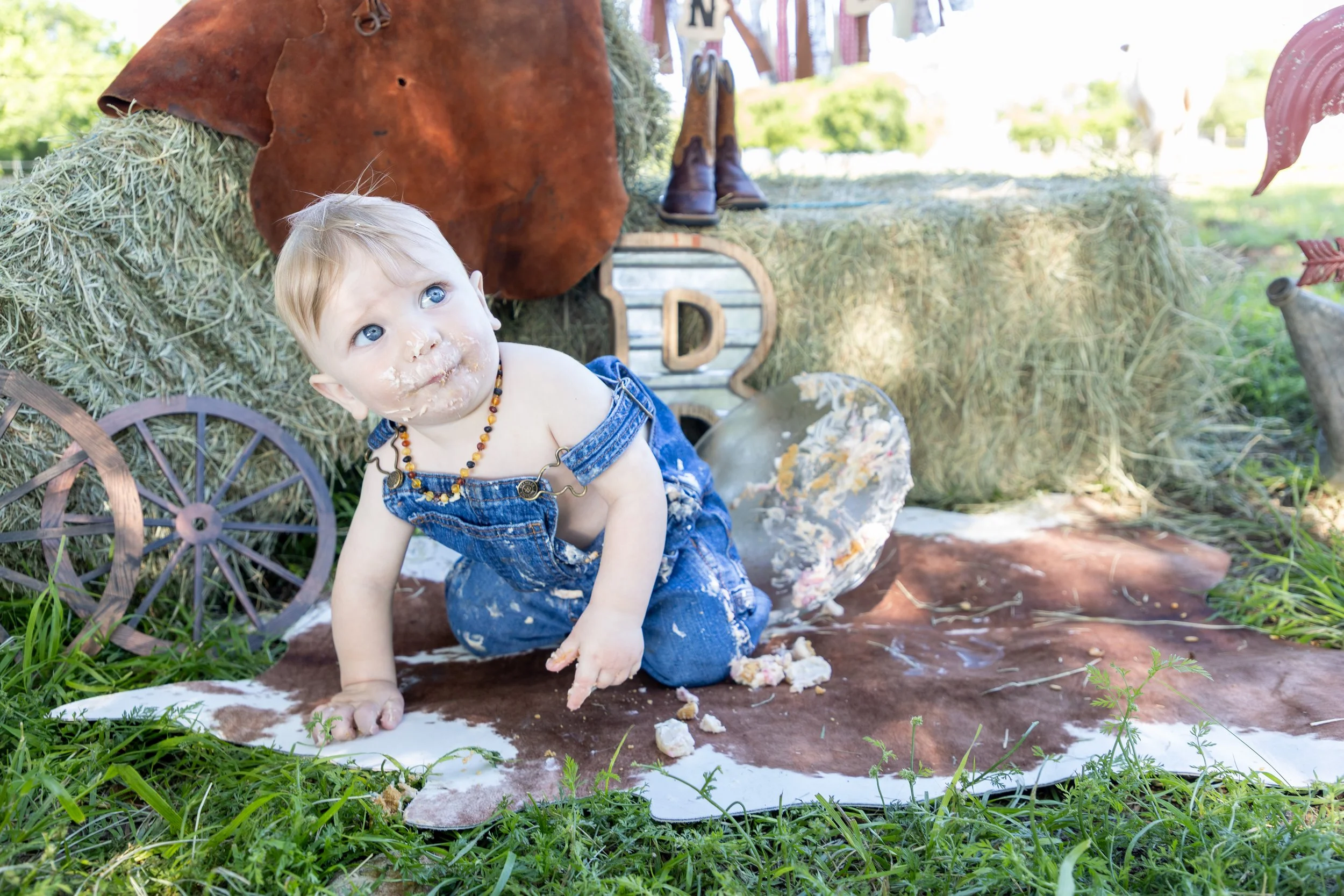 A young child with blond hair, blue eyes, and a necklace, wearing denim overalls, kneeling on the ground with food and cake crumbs on their face and hands. The child is outdoors on a cowhide rug with a festival or farm-themed setup behind them, inclu